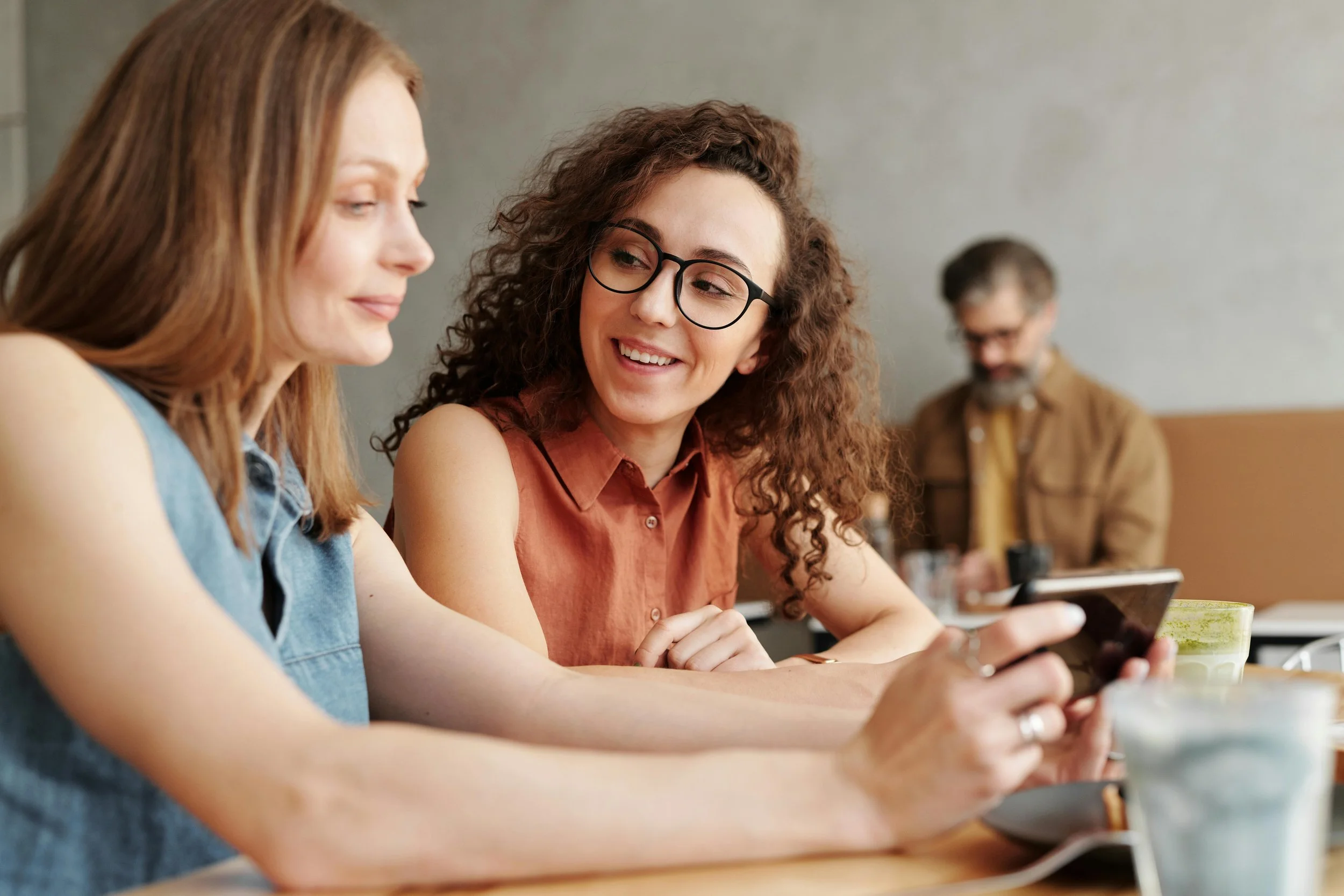 Two women sitting at a table in a cafe, smiling and looking at a smartphone together, with a man working on a laptop in the background.