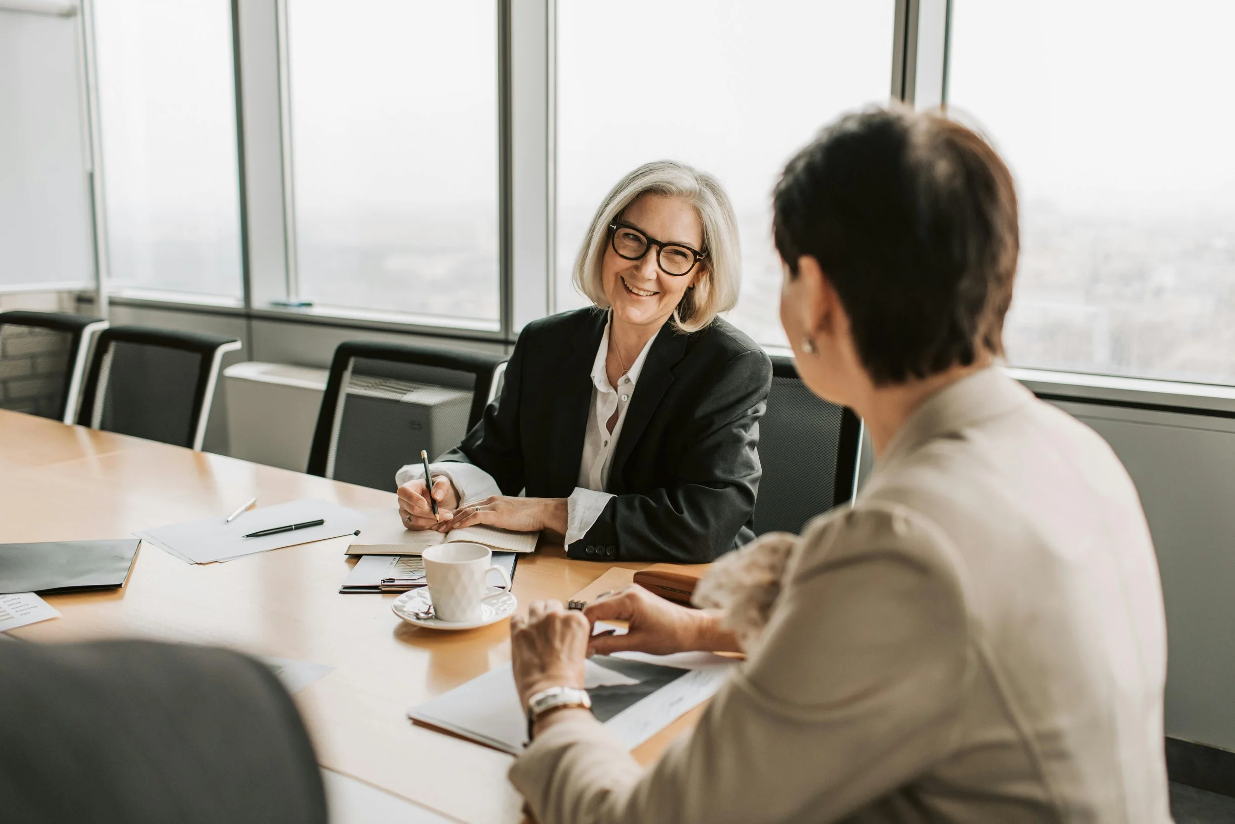 Two women having a business meeting in a conference room with large windows, one woman smiling and taking notes, the other woman holding papers.