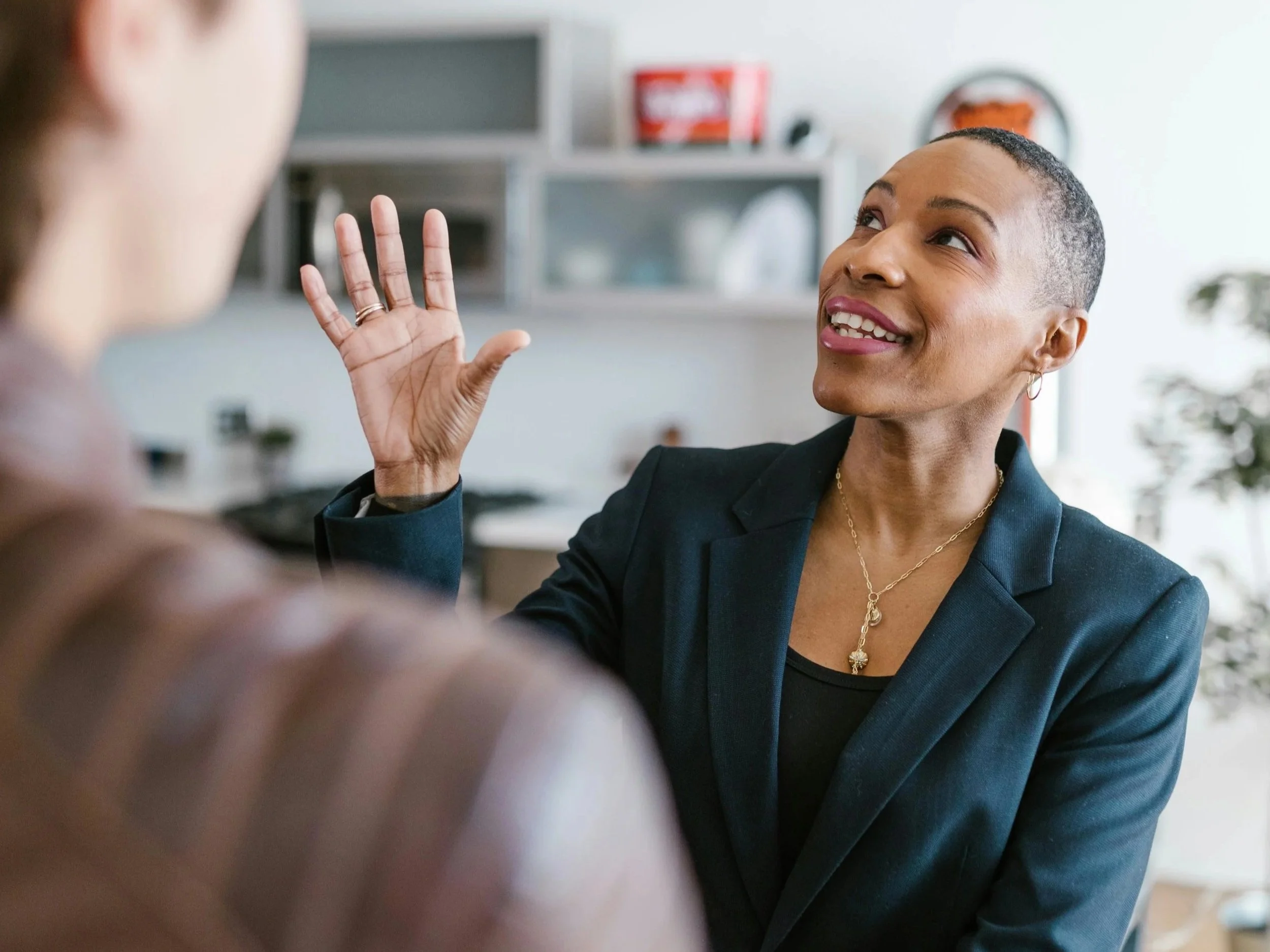 A woman in a blue blazer smiling and talking to another person with blurred shoulder in the foreground. She has short hair, wears earrings and a necklace, and gestures with her right hand.