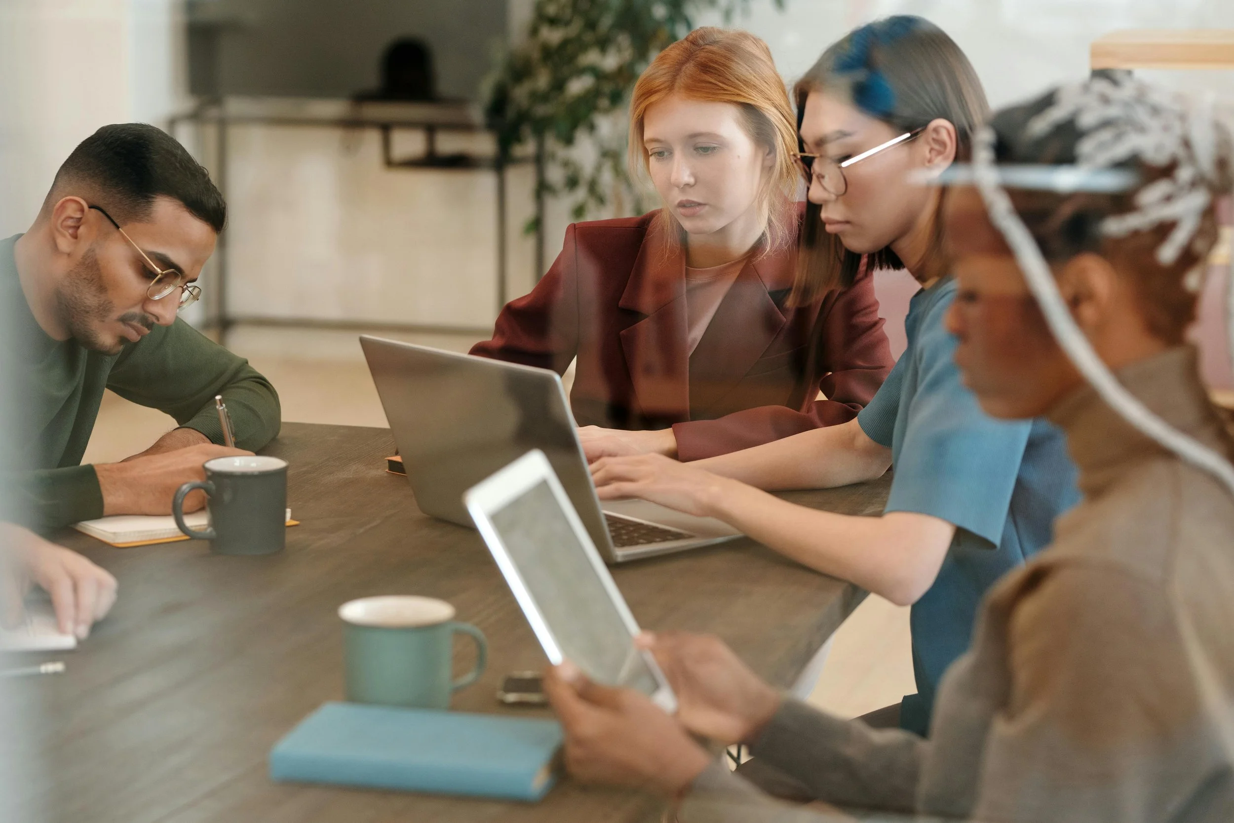 A diverse group of four young adults working together at a wooden table in a modern office. They are using laptops, tablets, and notebooks, with coffee mugs also on the table.