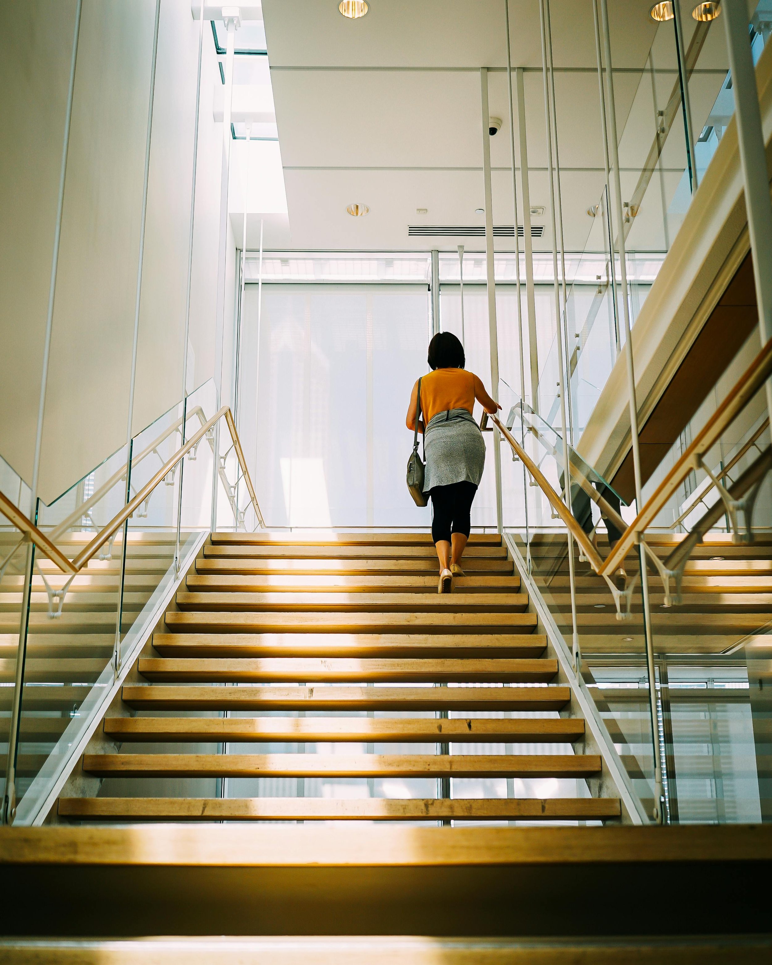 A woman in a yellow top, black leggings, and a gray sweater tied around her waist ascending a wooden staircase inside a modern building with glass walls and railings.