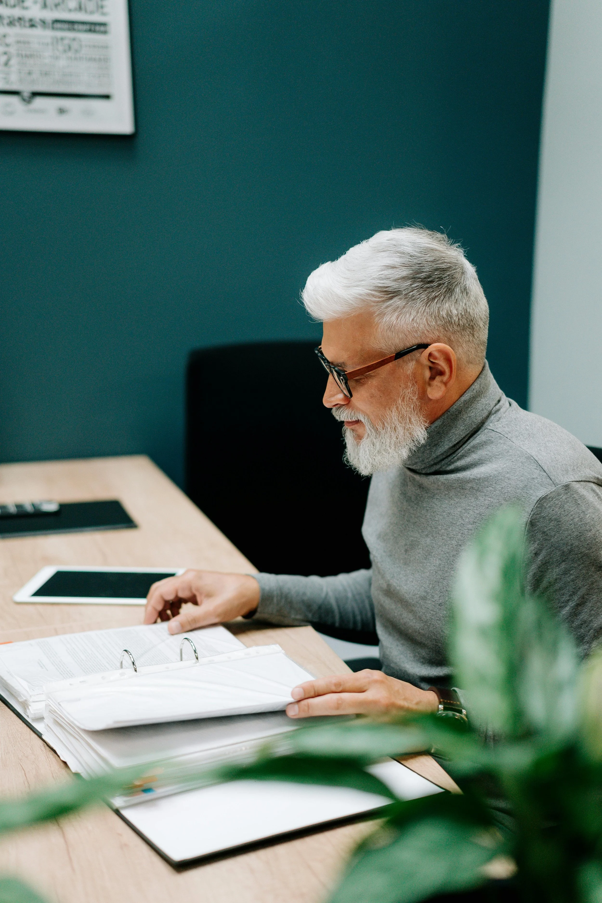 An elderly man with glasses and a white beard reading documents at a desk in an office.