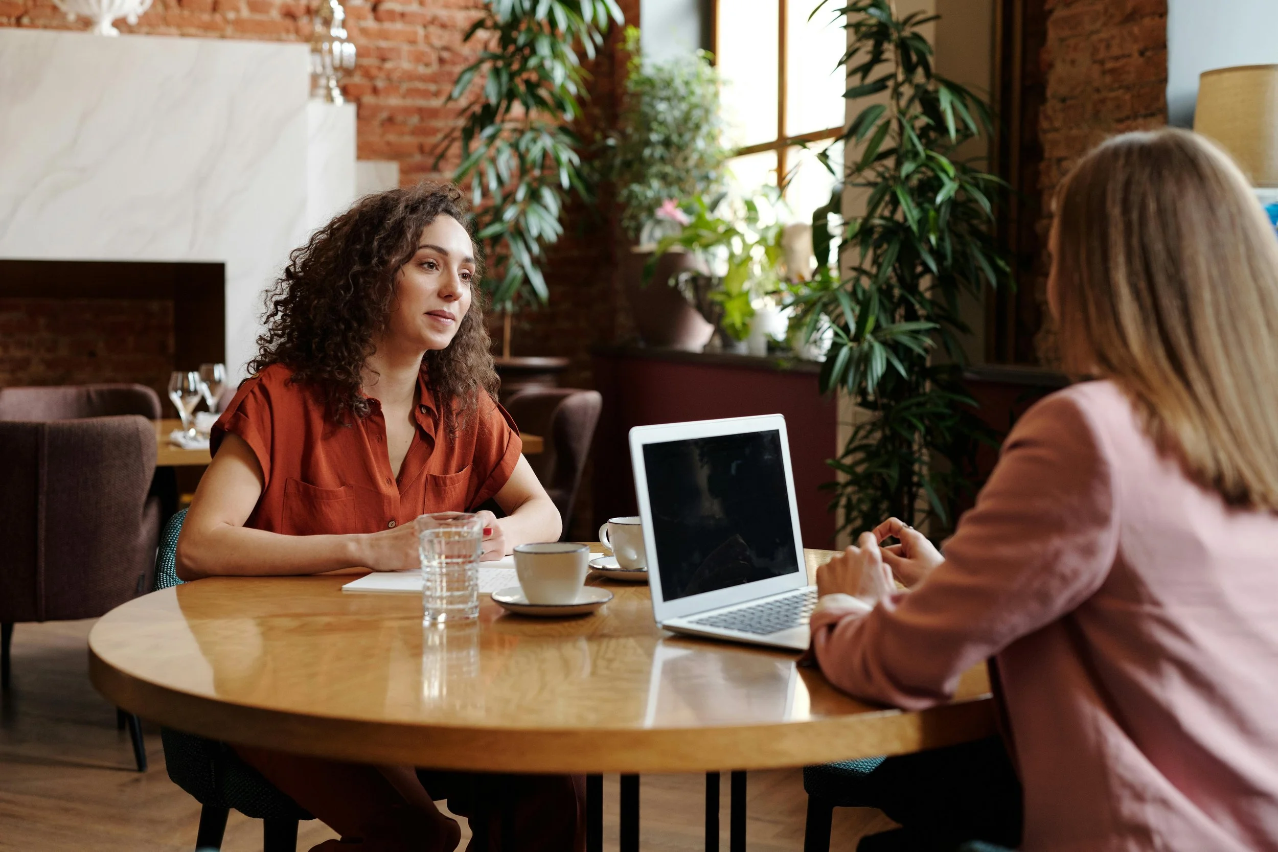 Two women having a conversation at a round wooden table in a cozy cafe or restaurant, with one woman facing the camera and the other with her back to the camera, a laptop, books, cups, and glasses on the table, and plants and a brick wall in the background.