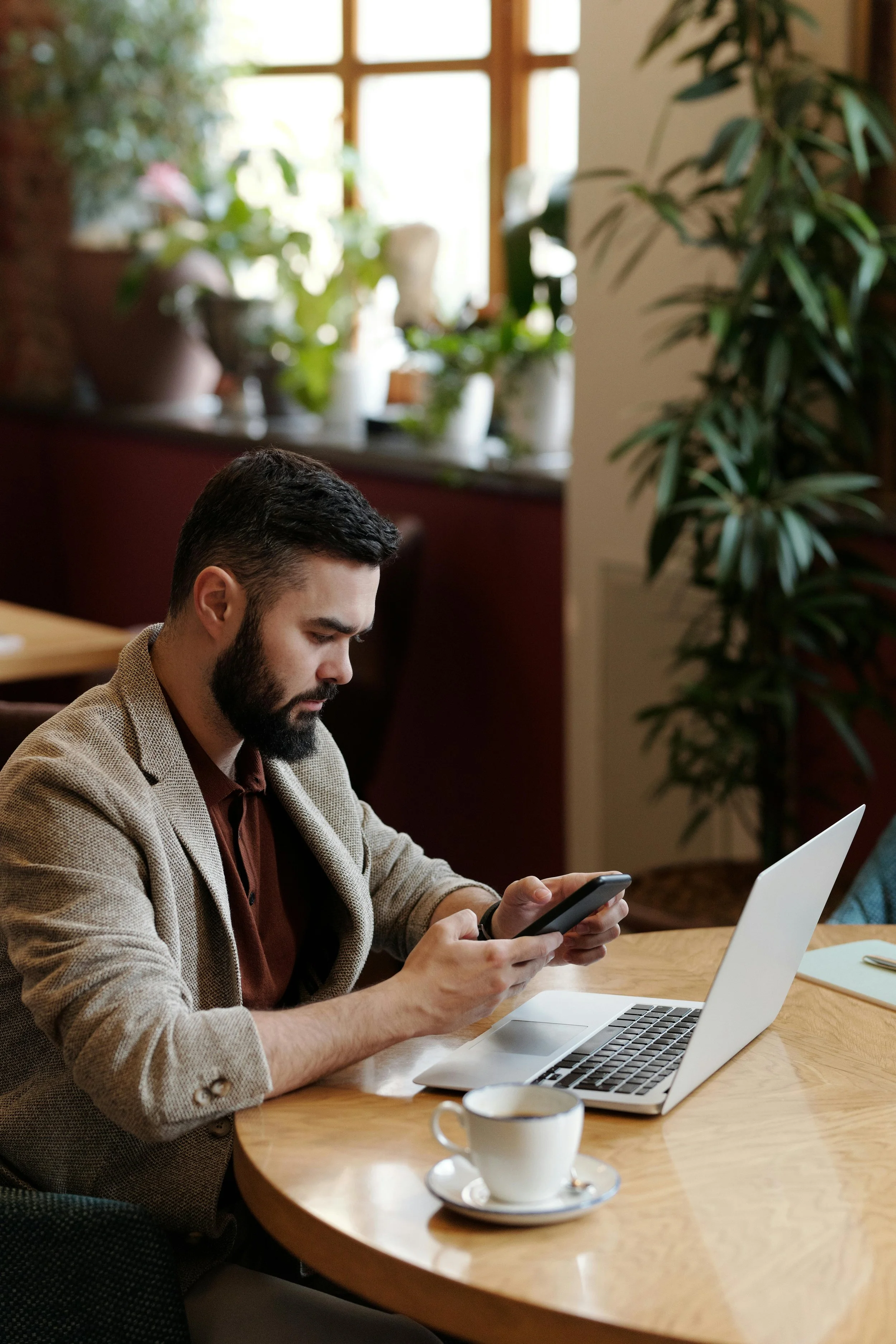 A man with a beard in a beige blazer and maroon shirt sitting at a wooden table with a silver laptop, holding a smartphone, with a cup of coffee on a saucer, in a cozy cafe with potted plants and a window in the background.