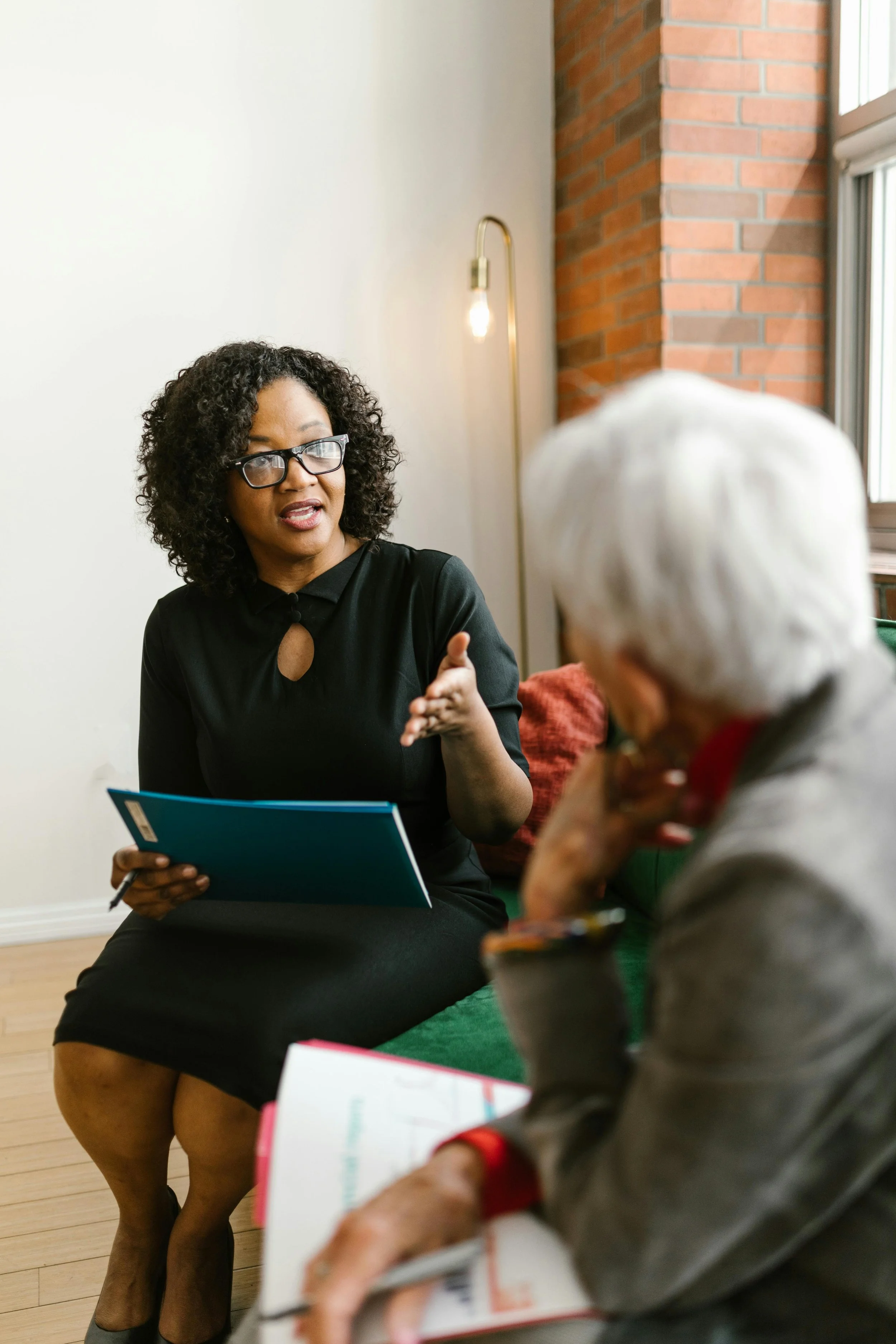 A woman with curly hair and glasses is speaking and holding a notebook, engaged in a discussion with an elderly woman with white hair who is taking notes.