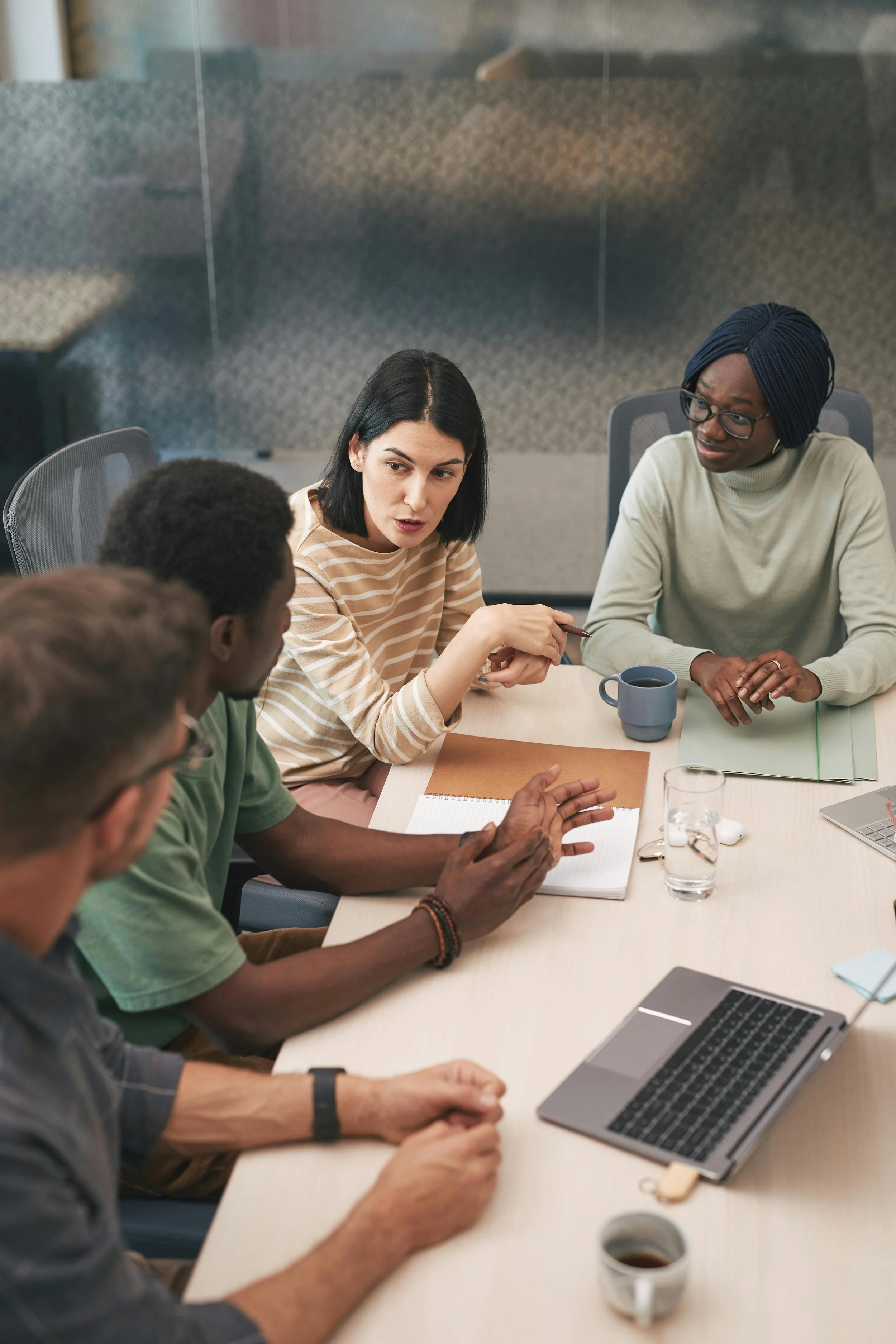Group of diverse people having a discussion around a conference table with laptops, notebooks, coffee cups, and glasses of water.