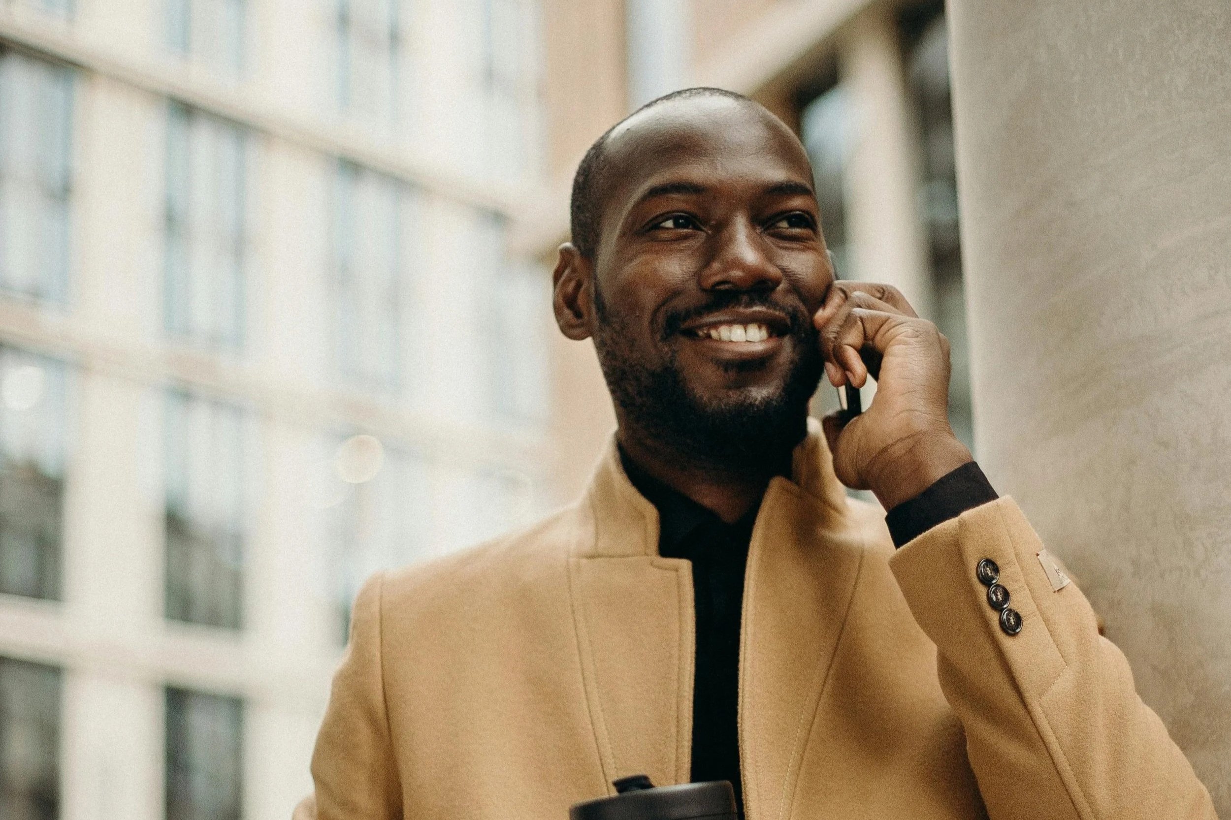 A smiling African American man in a tan coat talking on a cellphone outside in an urban setting.