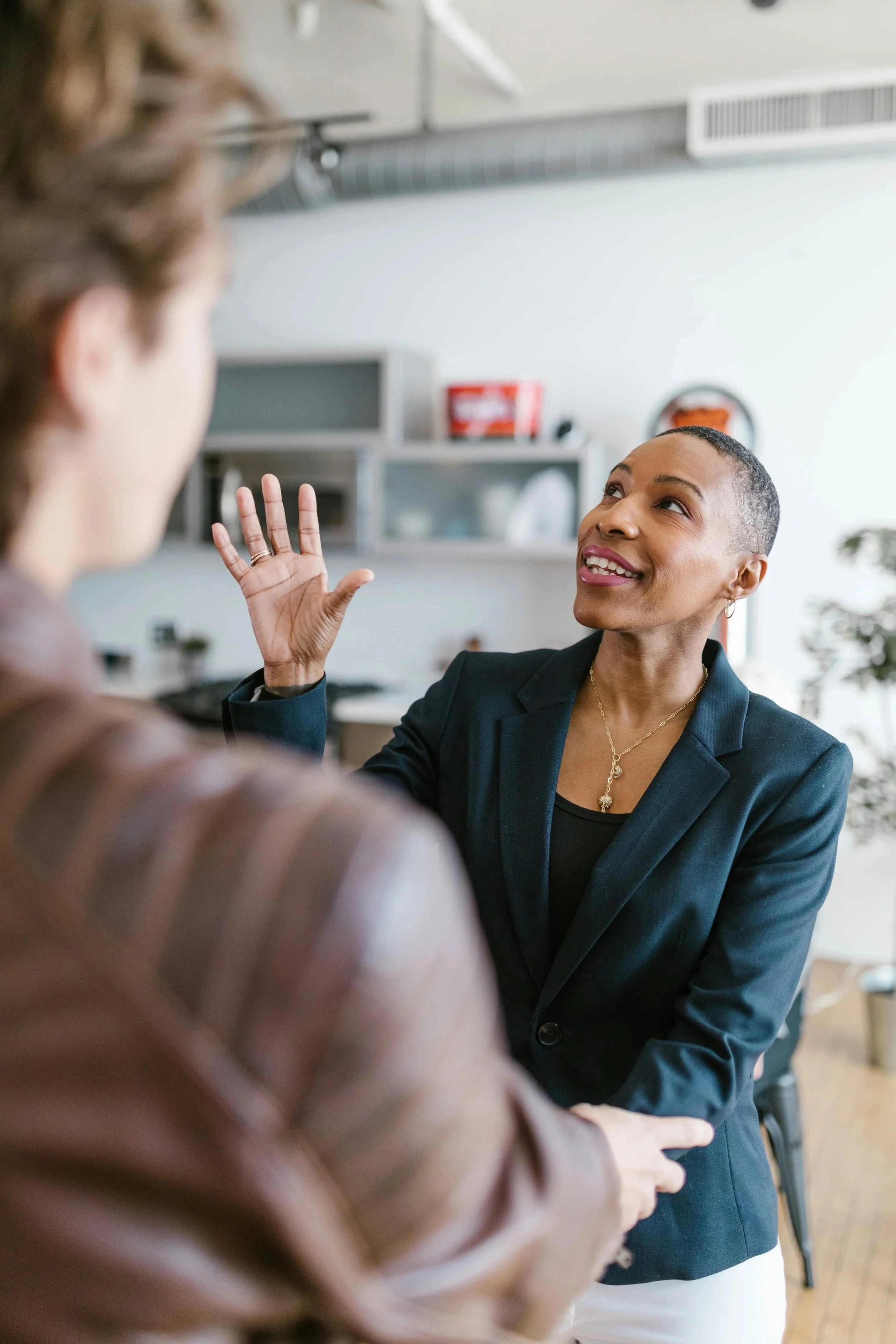 Two women engaging in conversation in a bright office setting, with one woman gesturing with her hand and smiling.