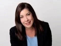 A woman with shoulder-length dark brown hair smiling at the camera, wearing a black blazer over a light blue shirt, against a plain white background.