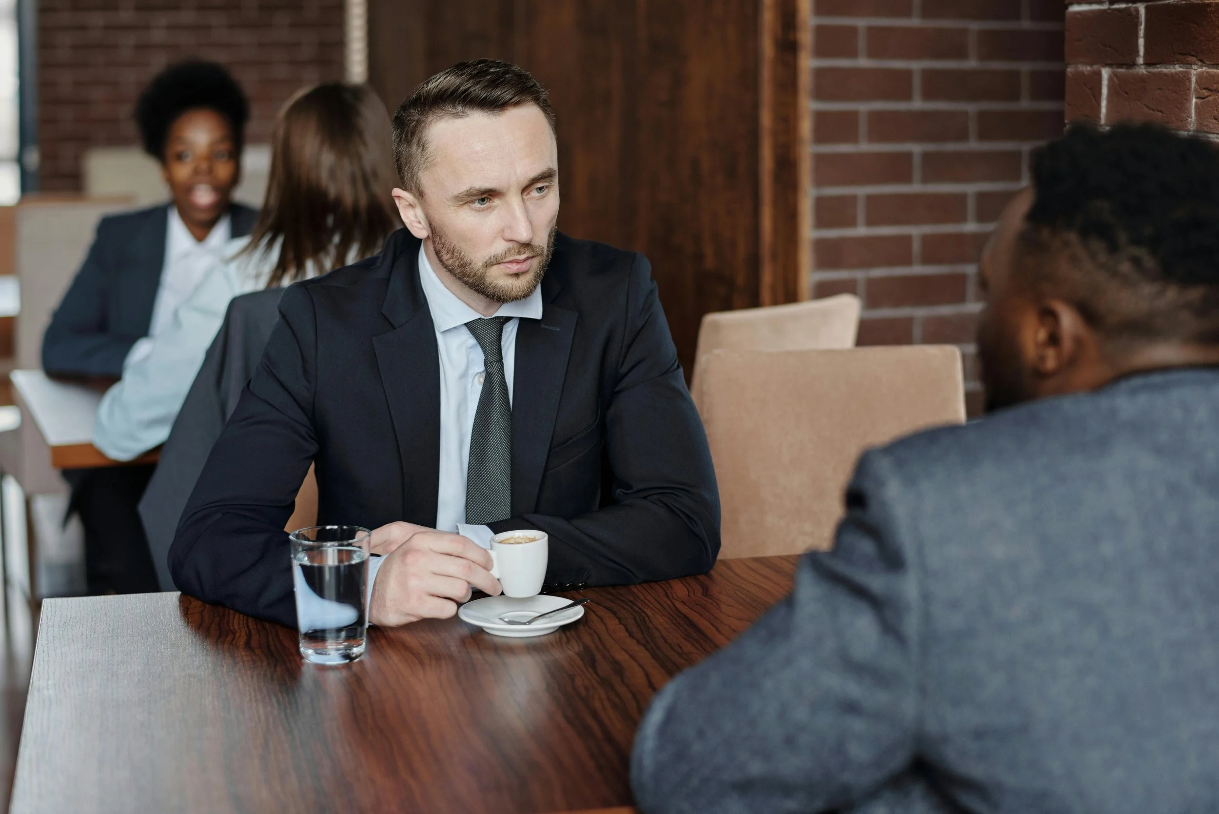 Two men are sitting at a wooden table in a coffee shop, engaged in a serious conversation. One man is holding a small cup of coffee, and there is a glass of water on the table. Two women are seated in the background, talking and smiling.