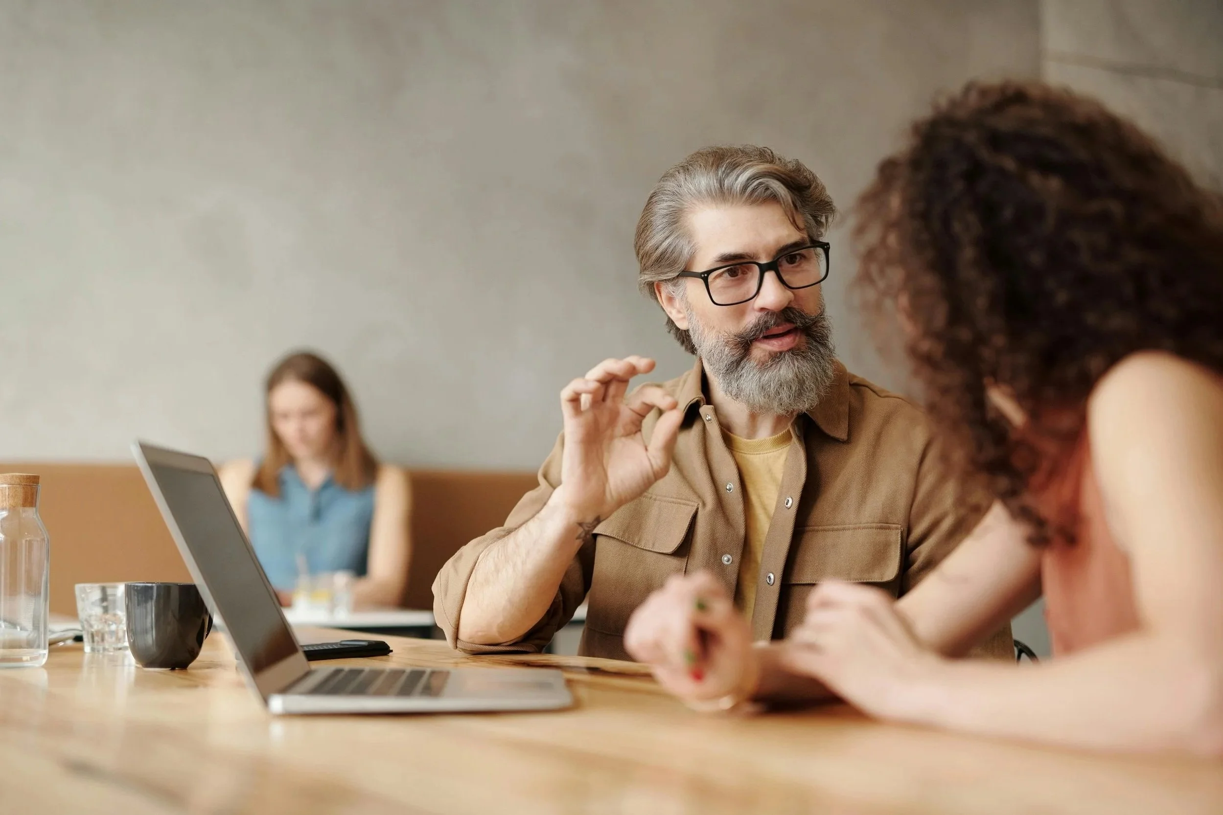A middle-aged man with glasses and gray hair, with a beard, wearing a brown shirt, is engaged in a conversation with a young woman with curly hair. They are seated at a wooden table with a laptop, a glass of water, and a cup. In the background, a woman with long hair is sitting at another table, smiling.