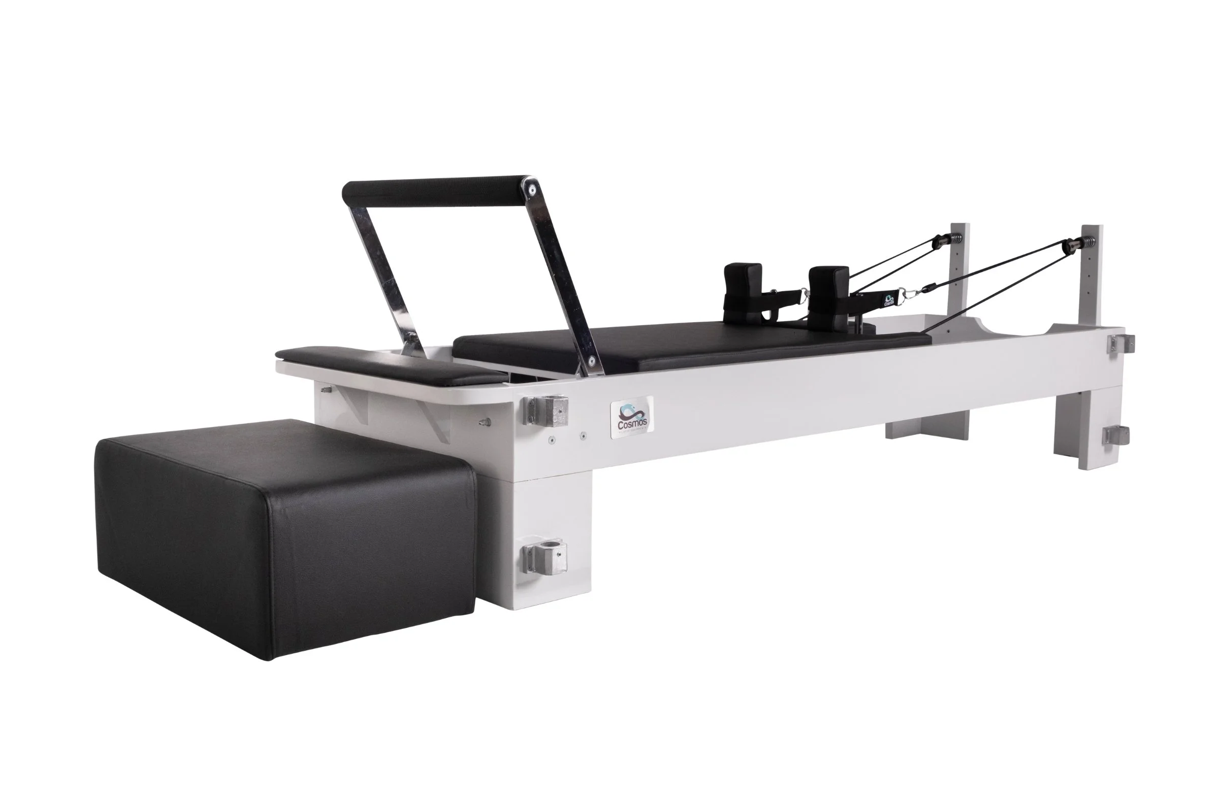 A Pilates reformer machine with a white frame, black padded platform, foot bar, and adjustable straps for resistance exercises.