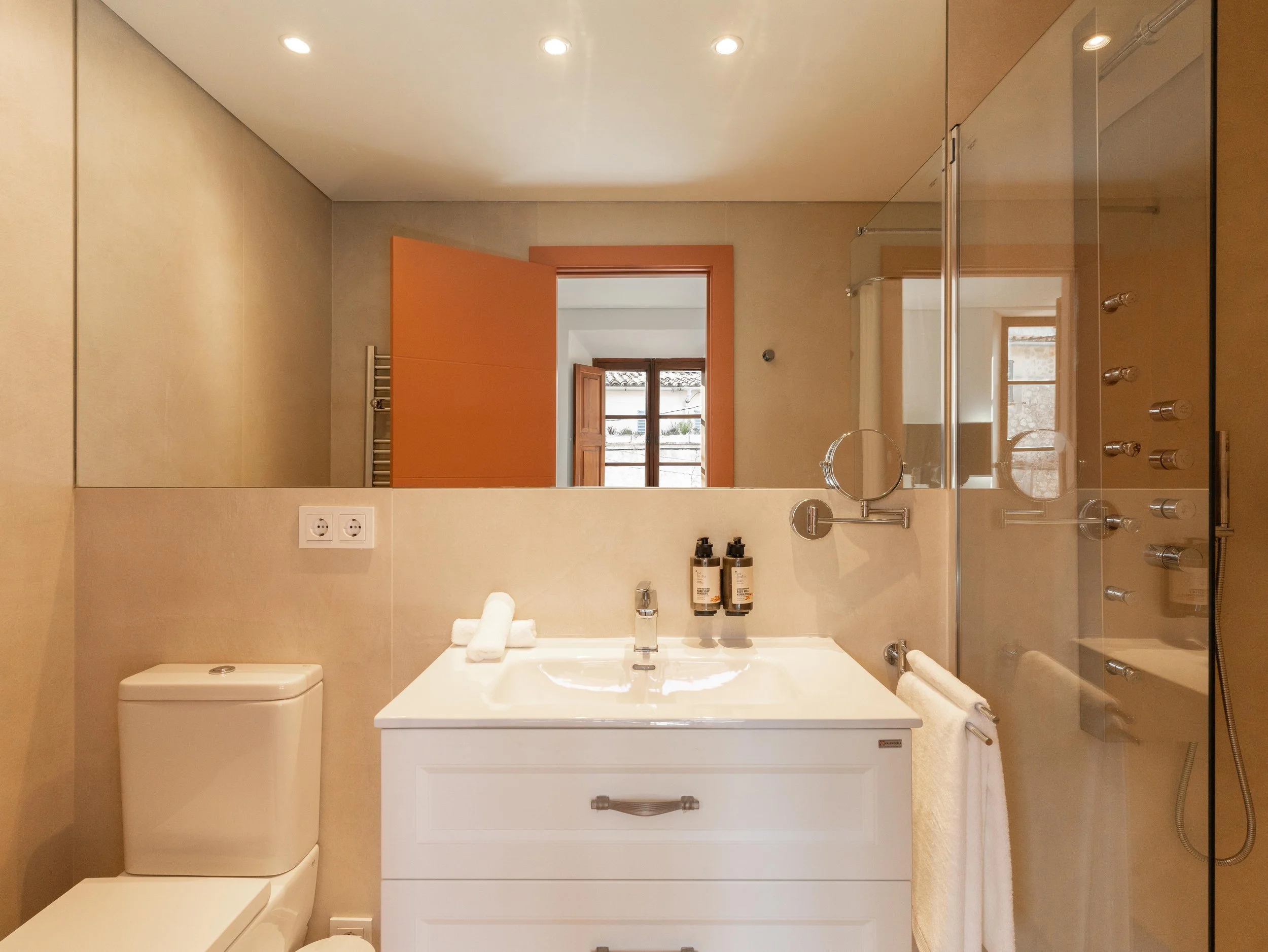 Baño con inodoro, lavabo con dos botellas de jabón, espejo grande, ducha con puerta de cristal y varias boquillas de hidromasaje, ventana y puerta de madera, toallas blancas, y enchufe doble en la pared.