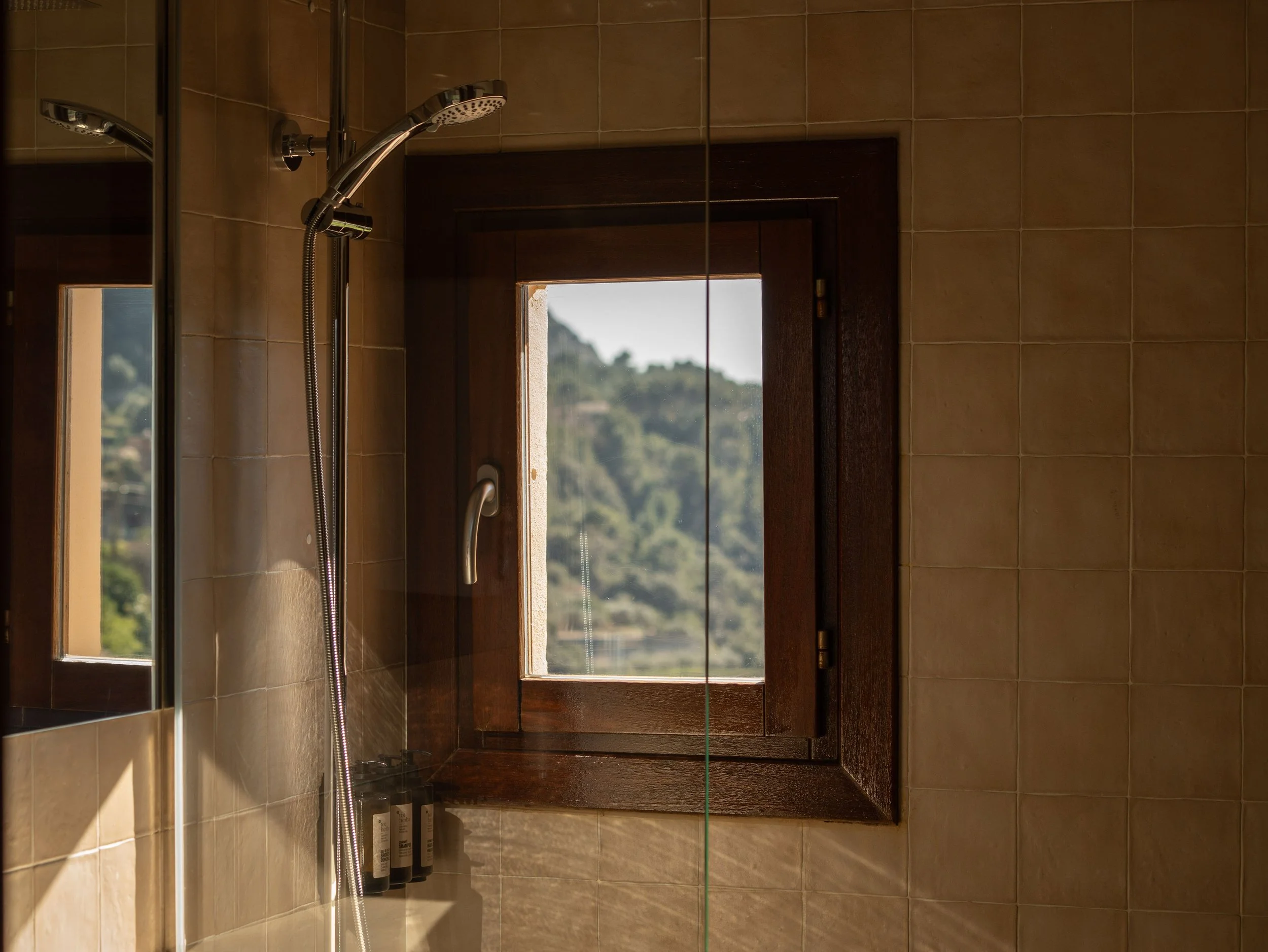 Baño con azulejos beige, ducha con cabezal y manguera, vista a un paisaje de colinas a través de una ventana de madera