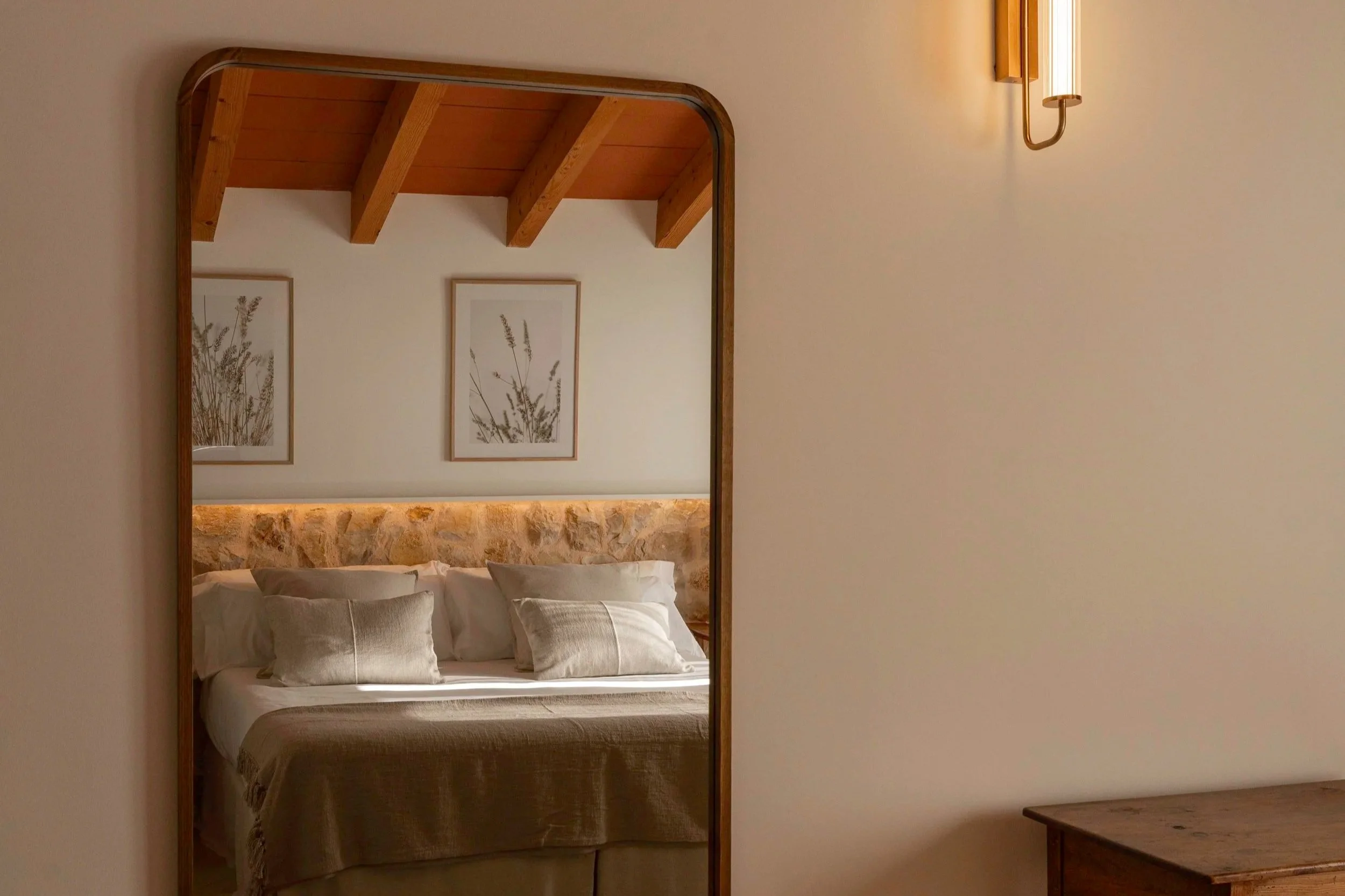 Espejo en pared que refleja una habitación con cama, cabecera de piedra, cuadros con plantas, paredes blancas y techo de madera con vigas.