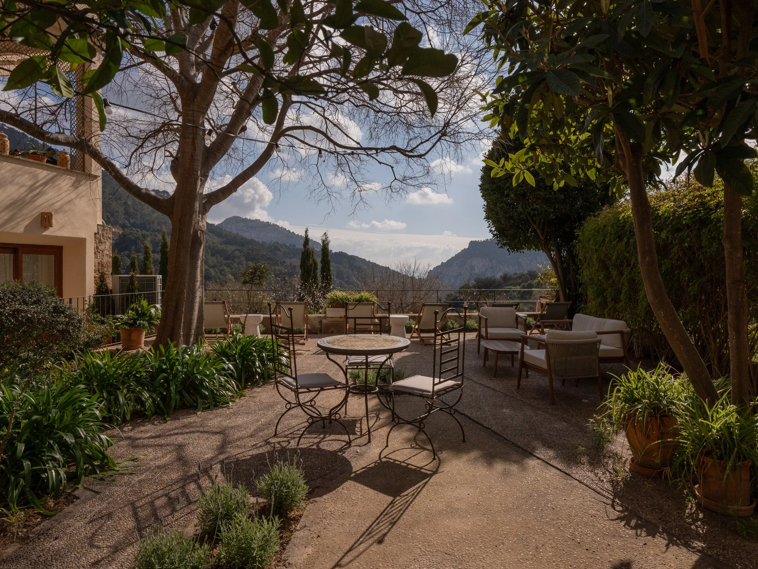 Un patio decorado con muebles de exterior, rodeado de plantas y árboles, con montañas y cielo con nubes en el fondo.