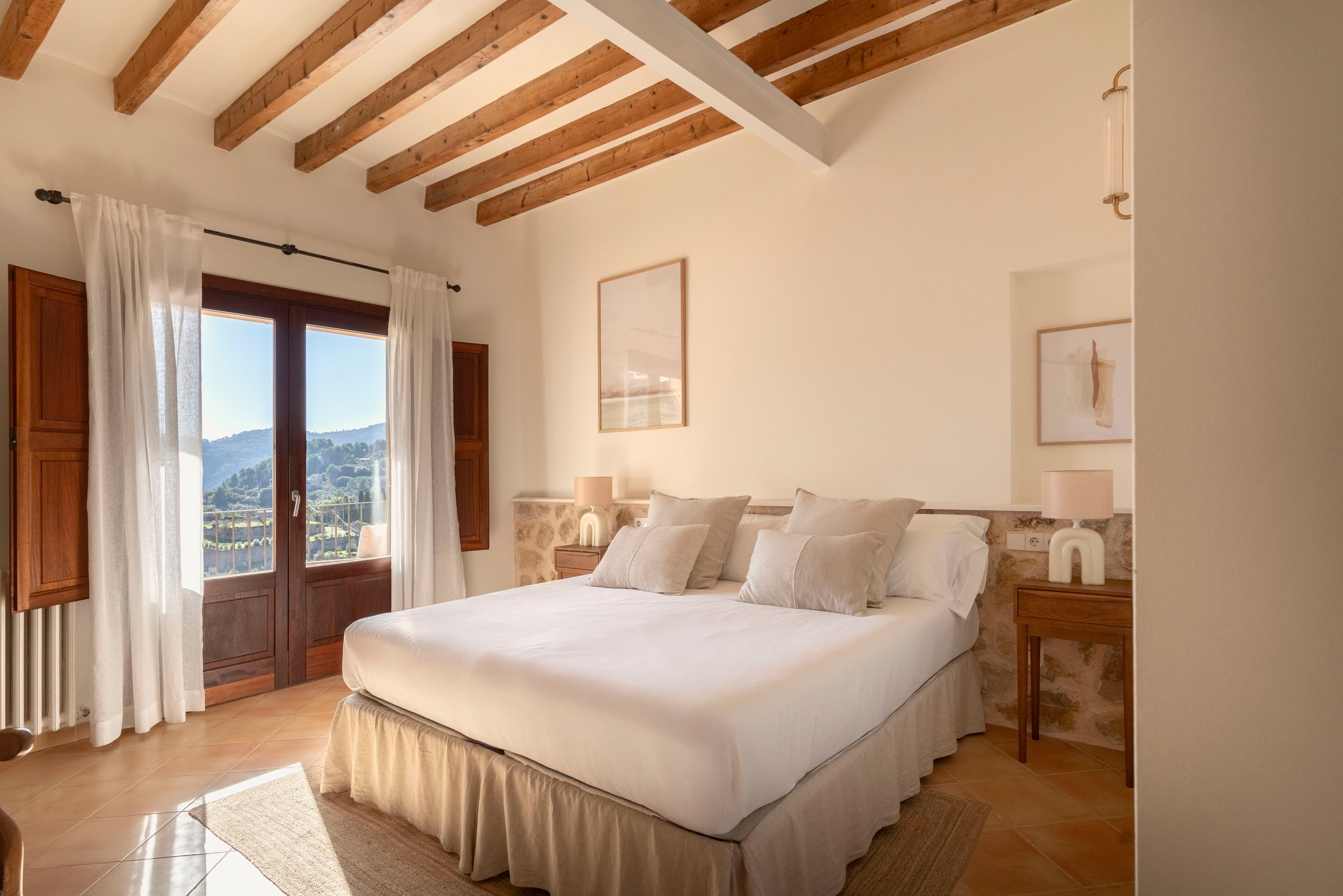 Habitación con cama grande, cortinas blancas, ventanas de madera con vista a montañas, paredes blancas, techo con vigas de madera, lámparas de noche a ambos lados de la cama, y cuadros decorativos.