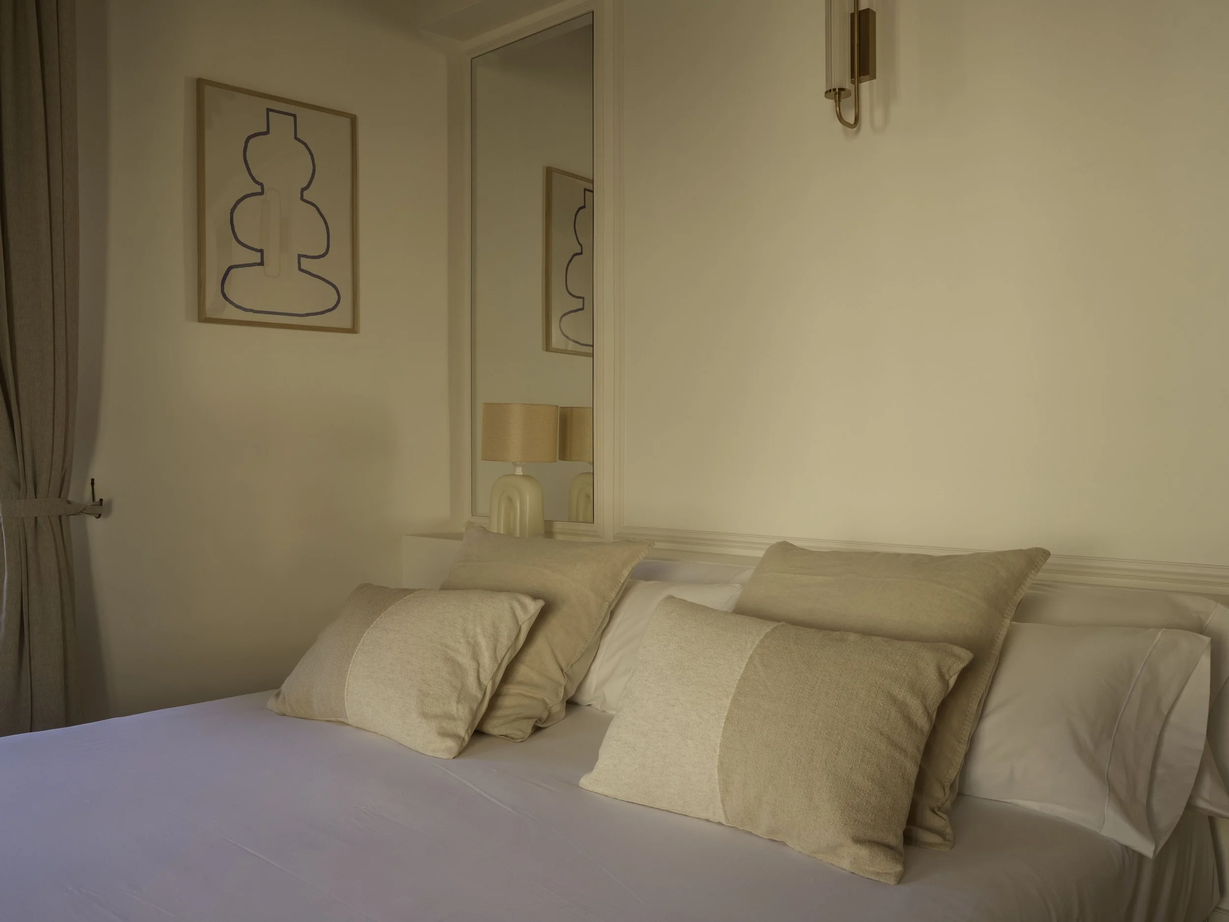 Cama con varias almohadas blancas y beige, en una habitación con paredes blancas, cuadro decorativo y lámpara de pared