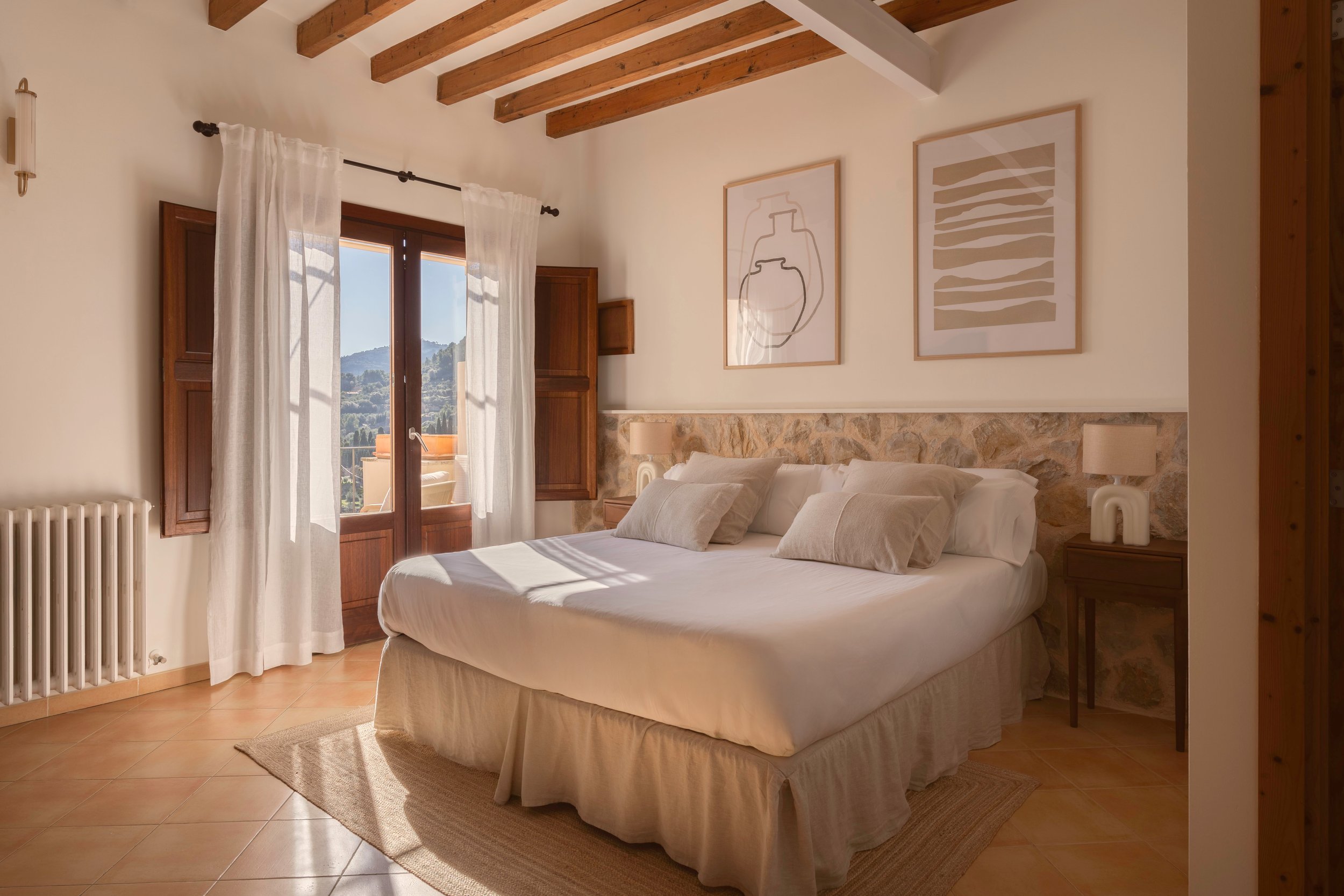 Habitación de estilo rústico con cama king size, cortinas blancas, pared de piedra, arte simple y vistas a las montañas.