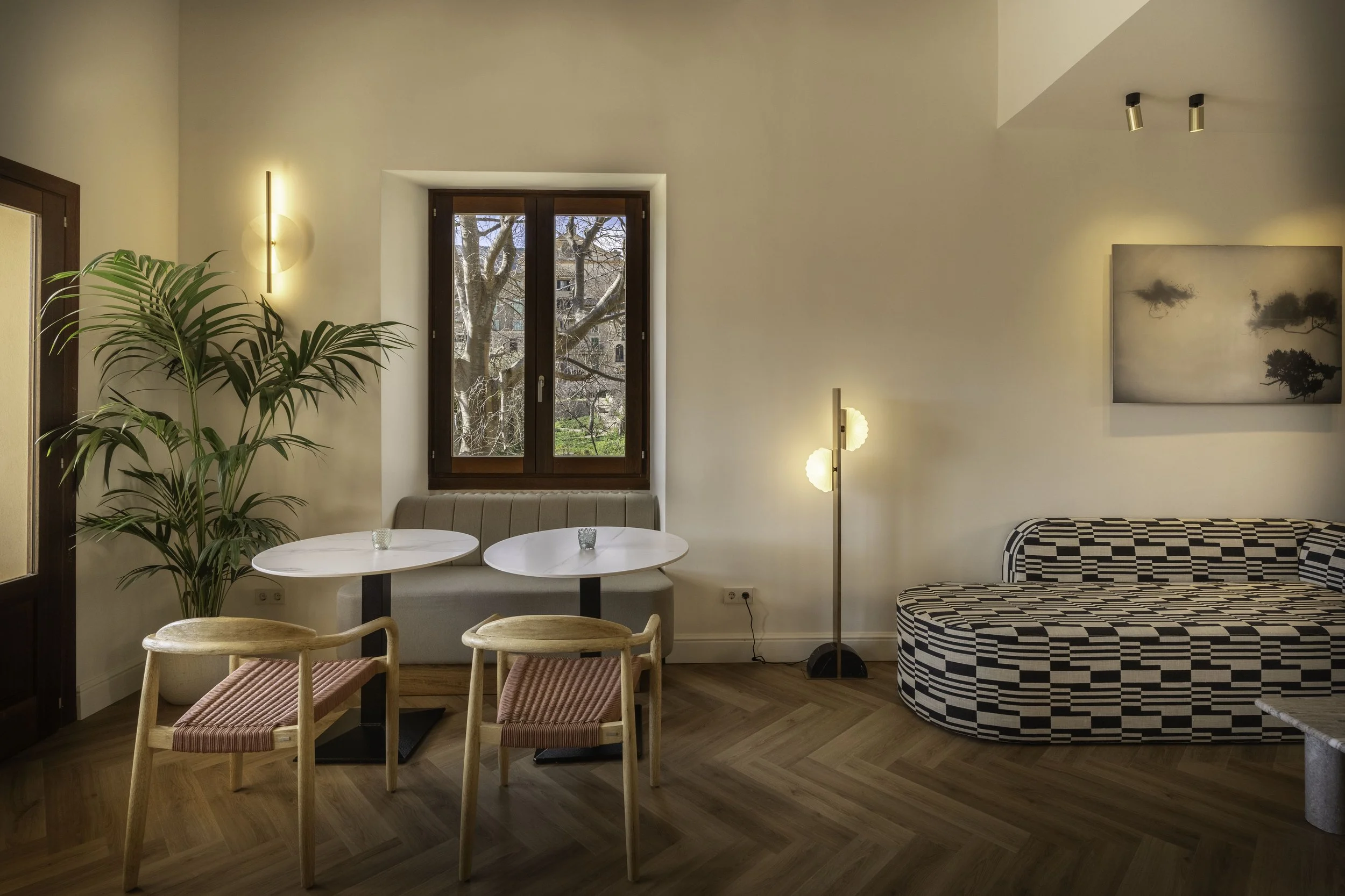 Sala de estar con un sillón curvado en negro y blanco, mesa de centro, una planta grande en la esquina, ventana con vista a árboles y edificios, lámpara de pie y pared con cuadros.