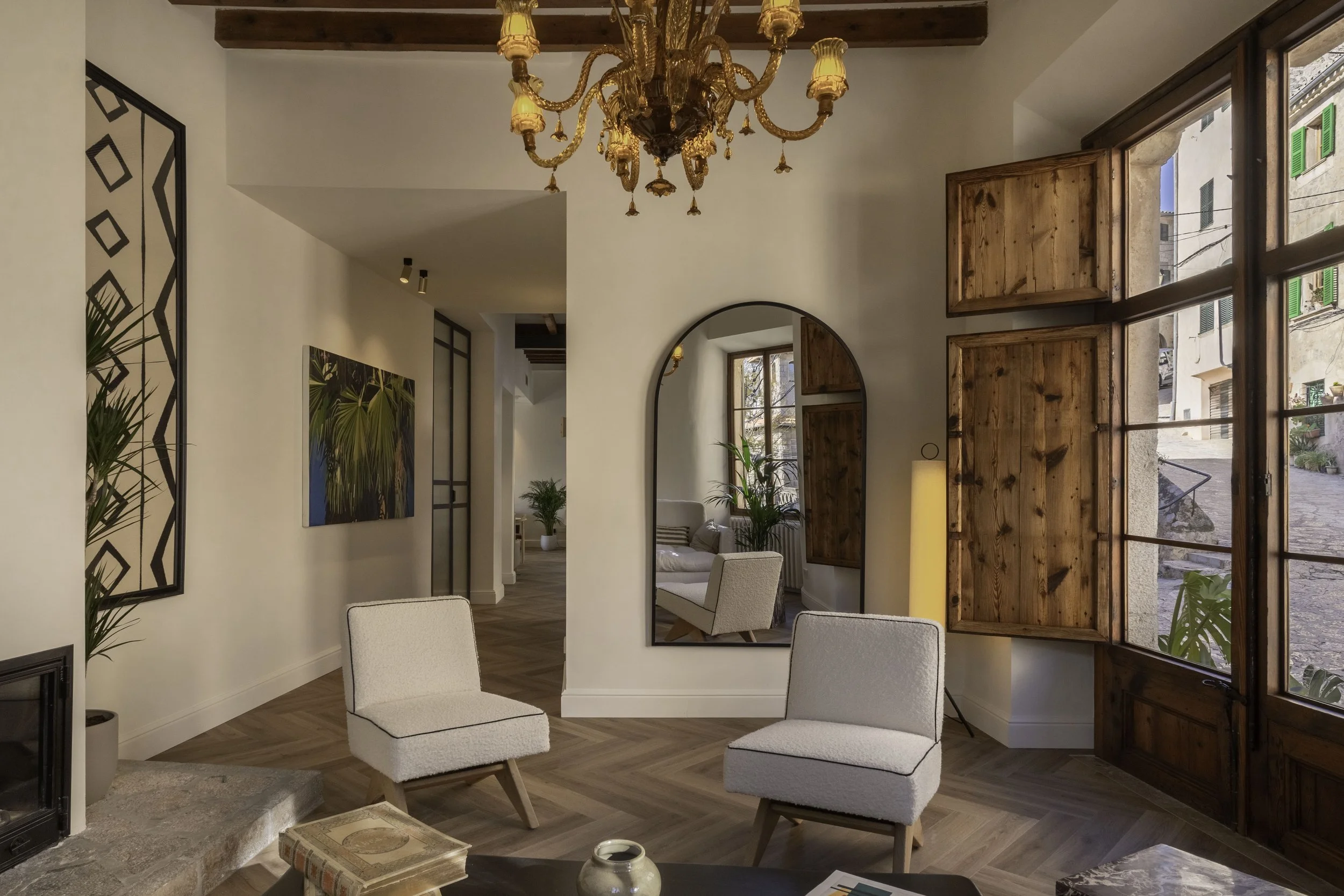 Sala de estar con silla beige, espejo de arco, ventanales de madera, plantas y lámpara de pie