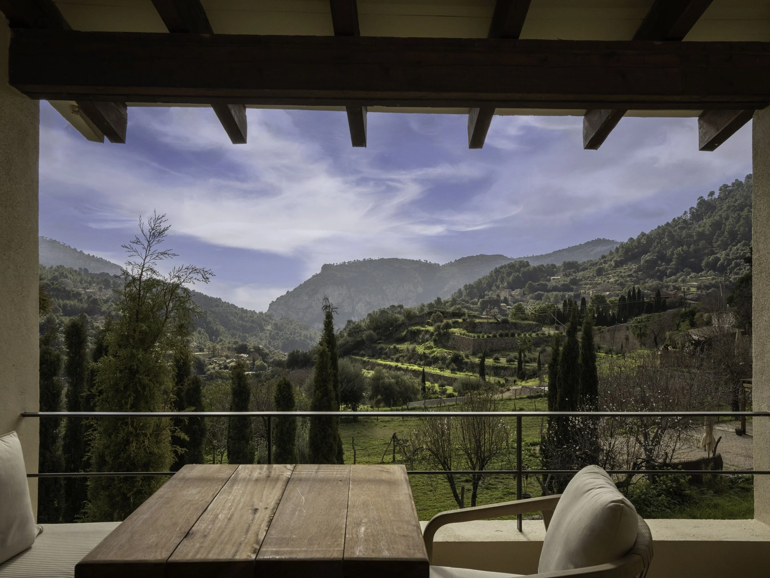 Vista de un paisaje montañoso con árboles y césped desde una terraza con mesa de madera y sillas, en un día soleado con cielo parcialmente nublado.