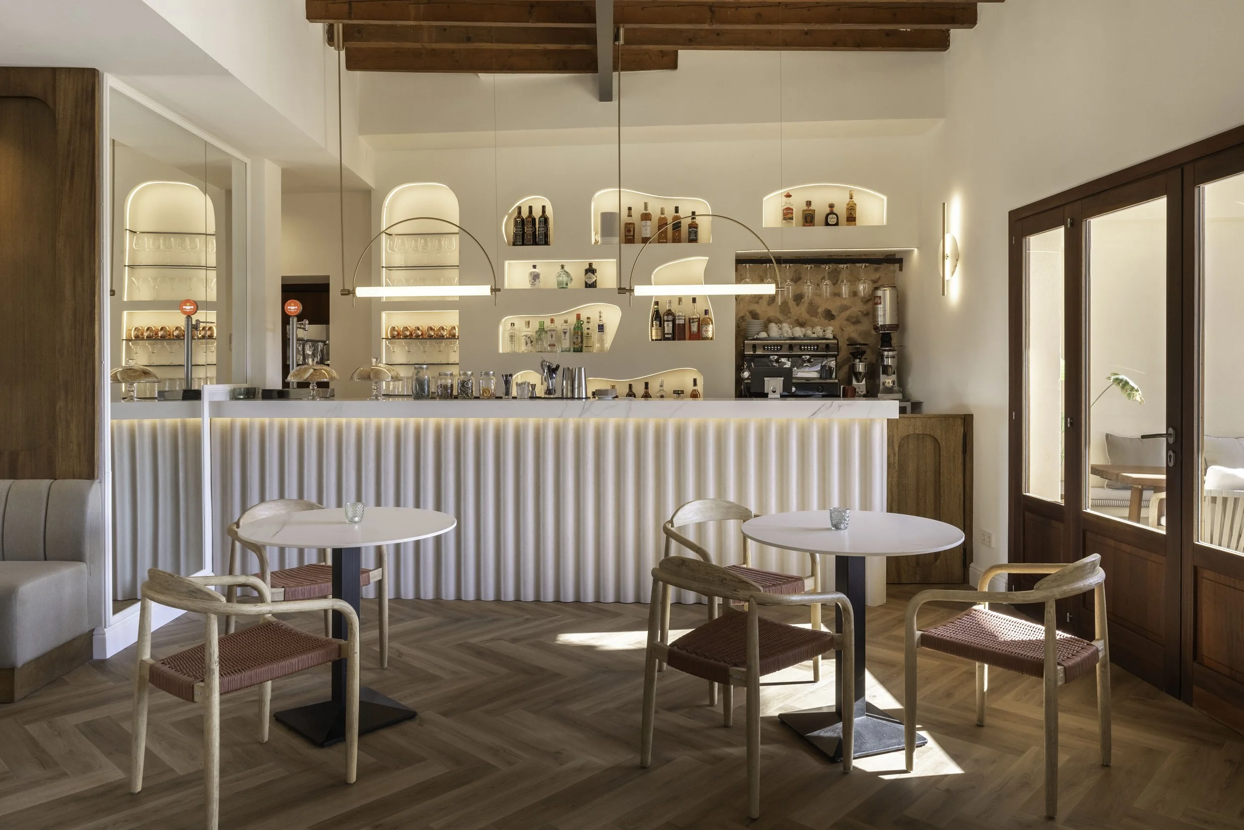 Interior de una cafetería moderna con mesas de madera y sillas de diseño minimalista, barra con iluminación suave y estanterías con botellas y utensilios, en un ambiente cálido y iluminado por luz natural.