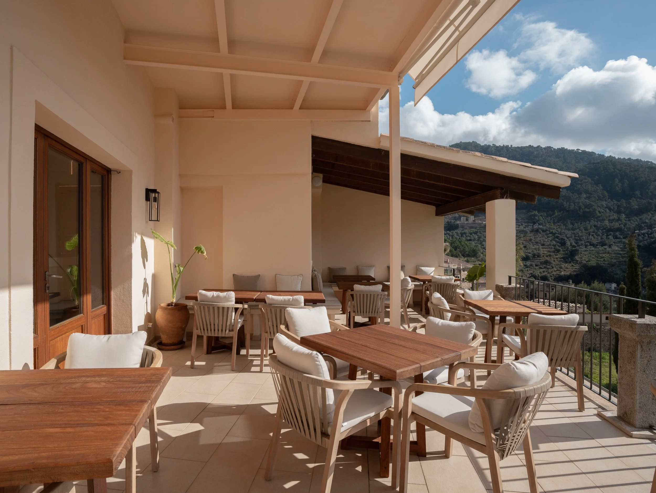 Terraza con mesas de madera, sillas con cojines blancos, plantas en macetas, vista de montañas y cielo con nubes.