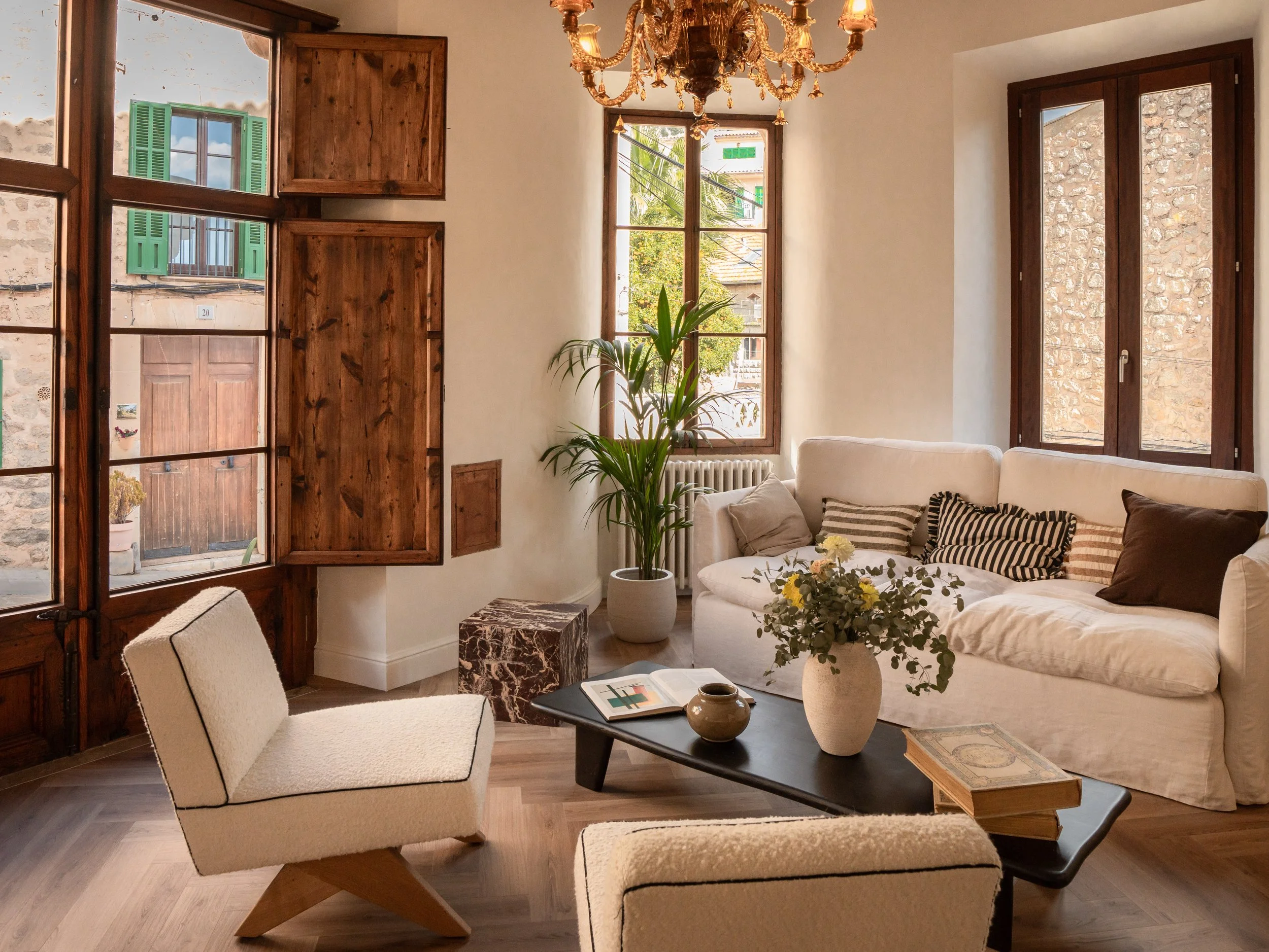 Sala de estar con sofa blanco, dos sillones, mesa de centro con libros y jarrón con flores, ventanas de madera, plantas y lámpara de araña.