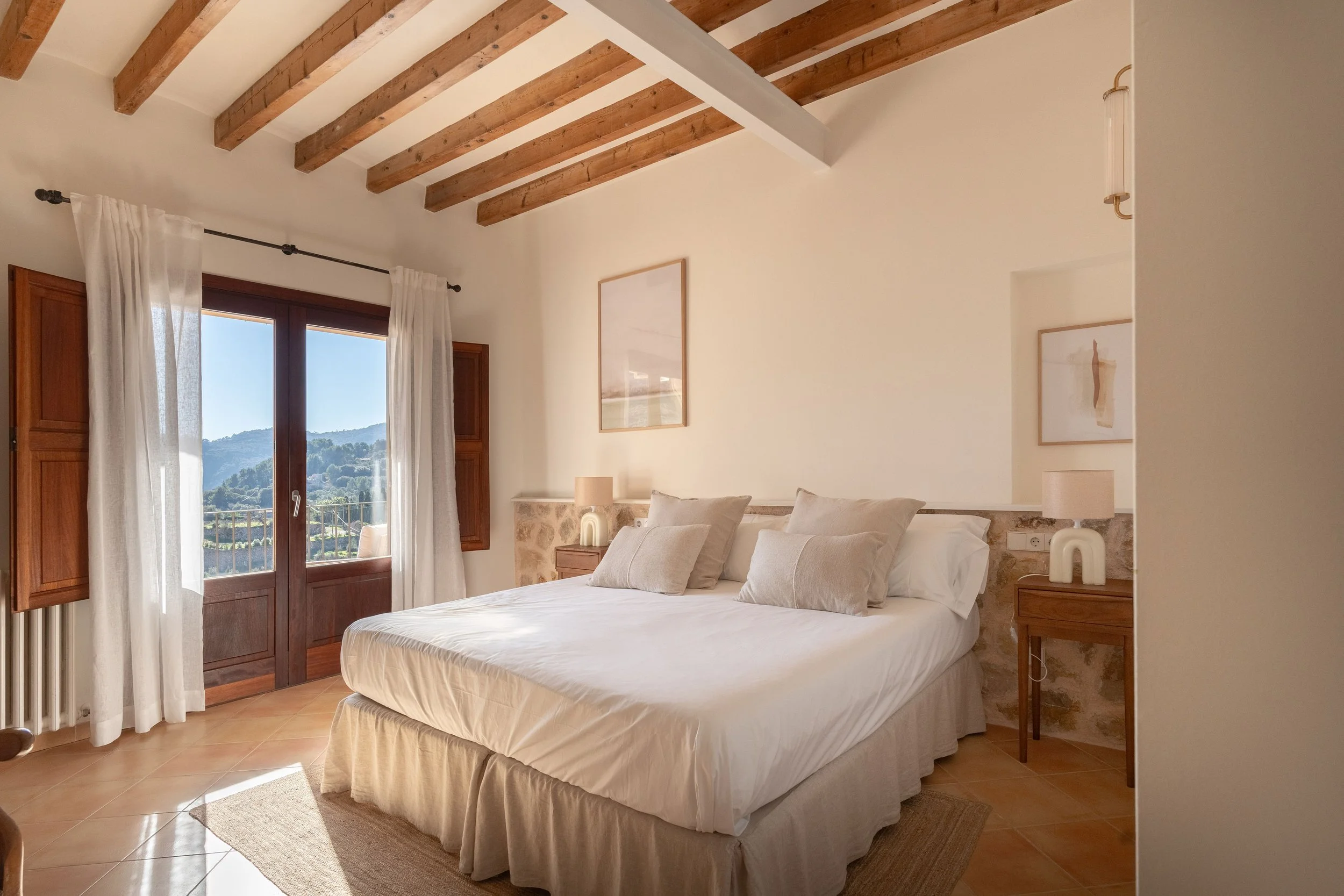 Habitación con cama grande, almohadas blancas, ventanas con cortinas blancas y vista hacia colinas verdes.