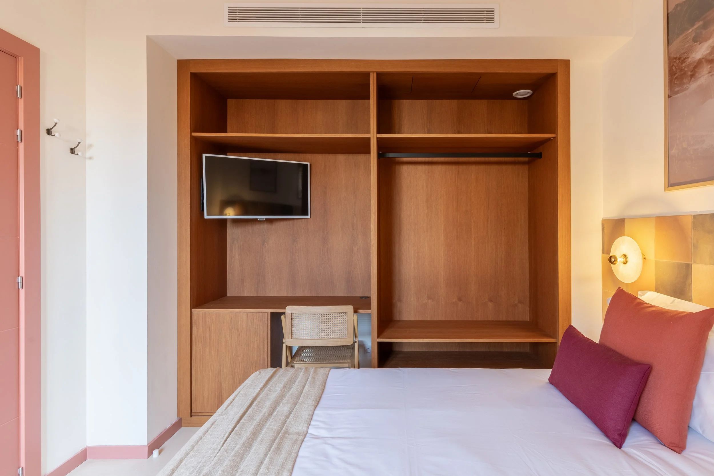 Habitación de hotel con cama, almohadas, lámpara y mesa con silla, armario de madera con televisor y estantes.