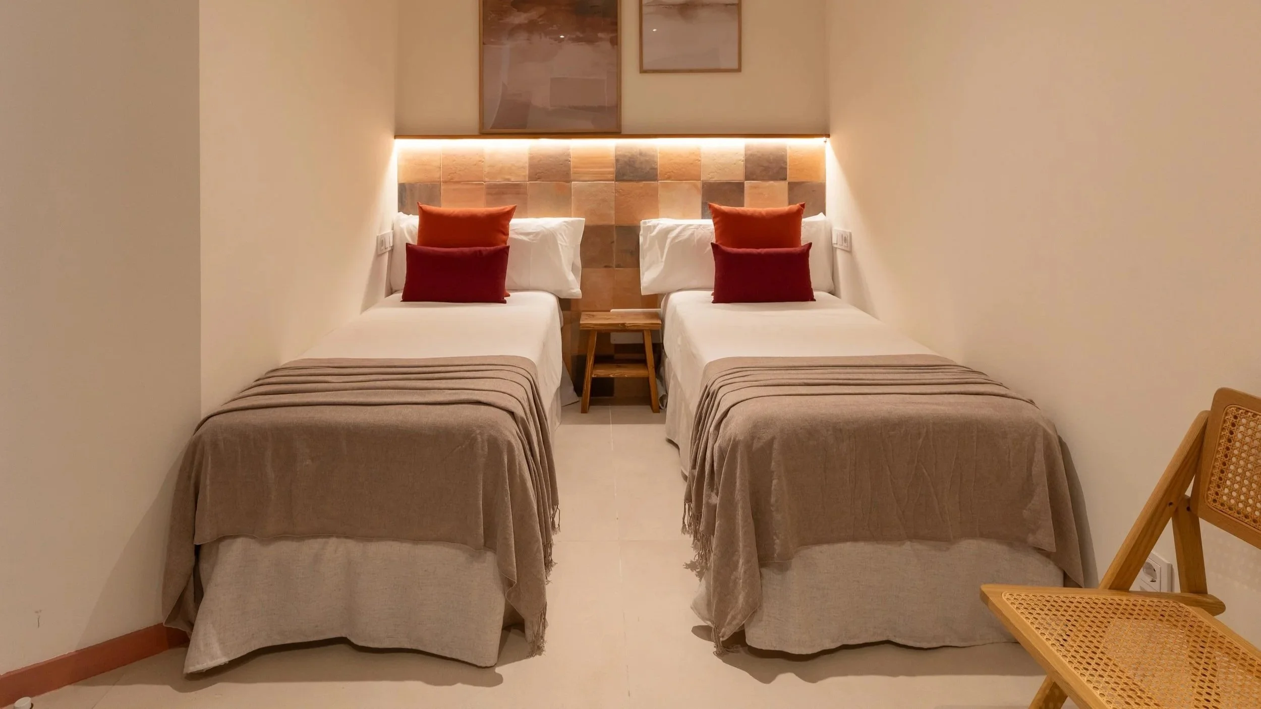 Dos camas individuales con cabeceras de azulejos de colores cálidos, cada una con almohadas blancas y cojines naranjas y rojos, en una habitación con paredes claras y una silla de madera a la derecha.