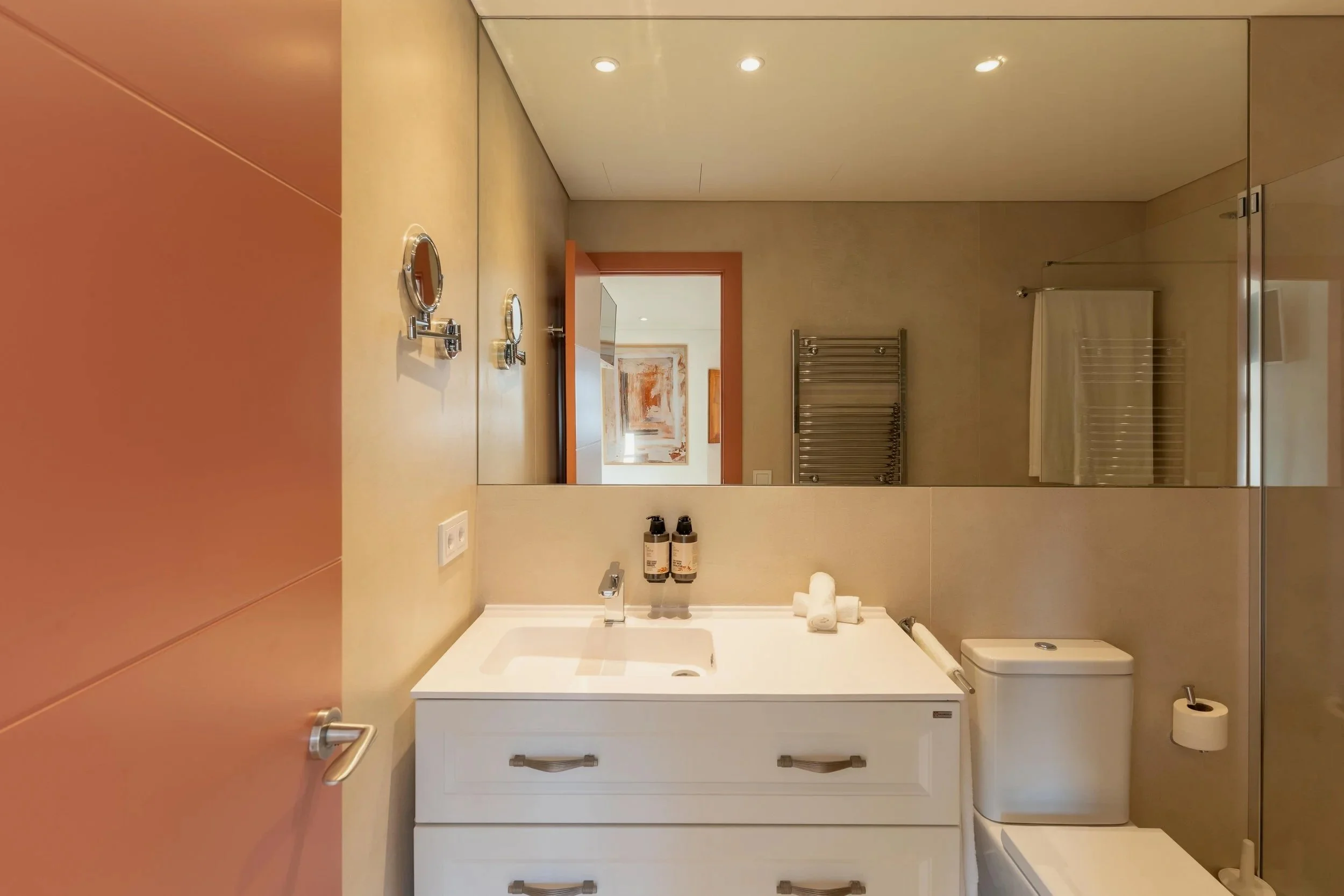 Baño con pared de azulejo beige, espejo grande, lavabo, toallas, jabón, inodoro y puerta de ducha con cristal