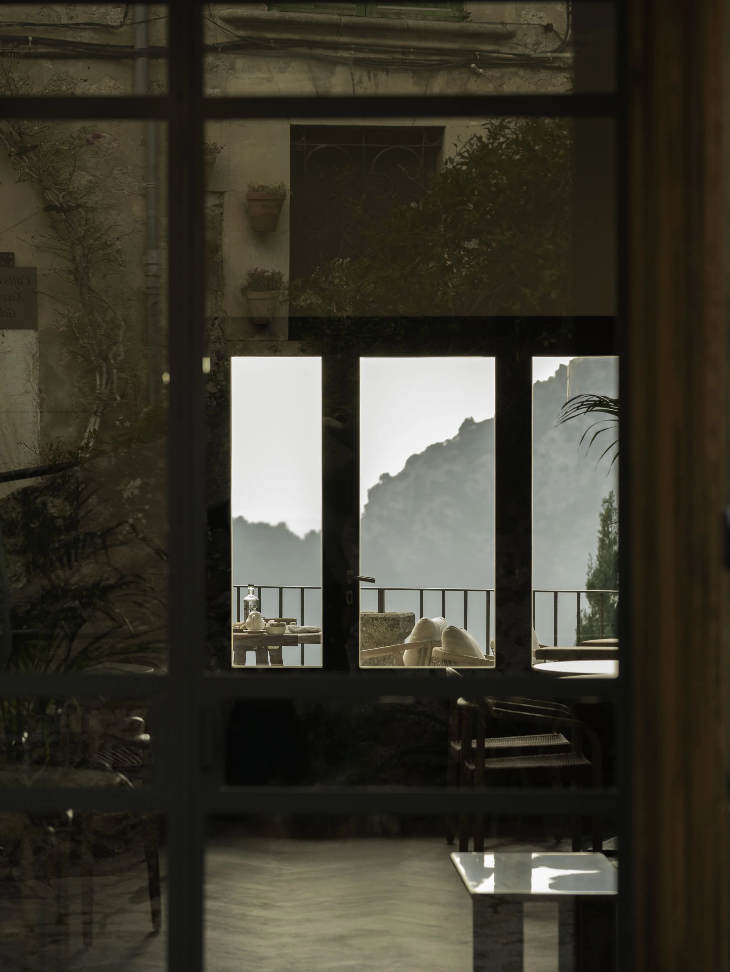 Vista de una terraza con sillas y mesa, vista a montañas en la distancia, vista a través de una ventana con persianas.