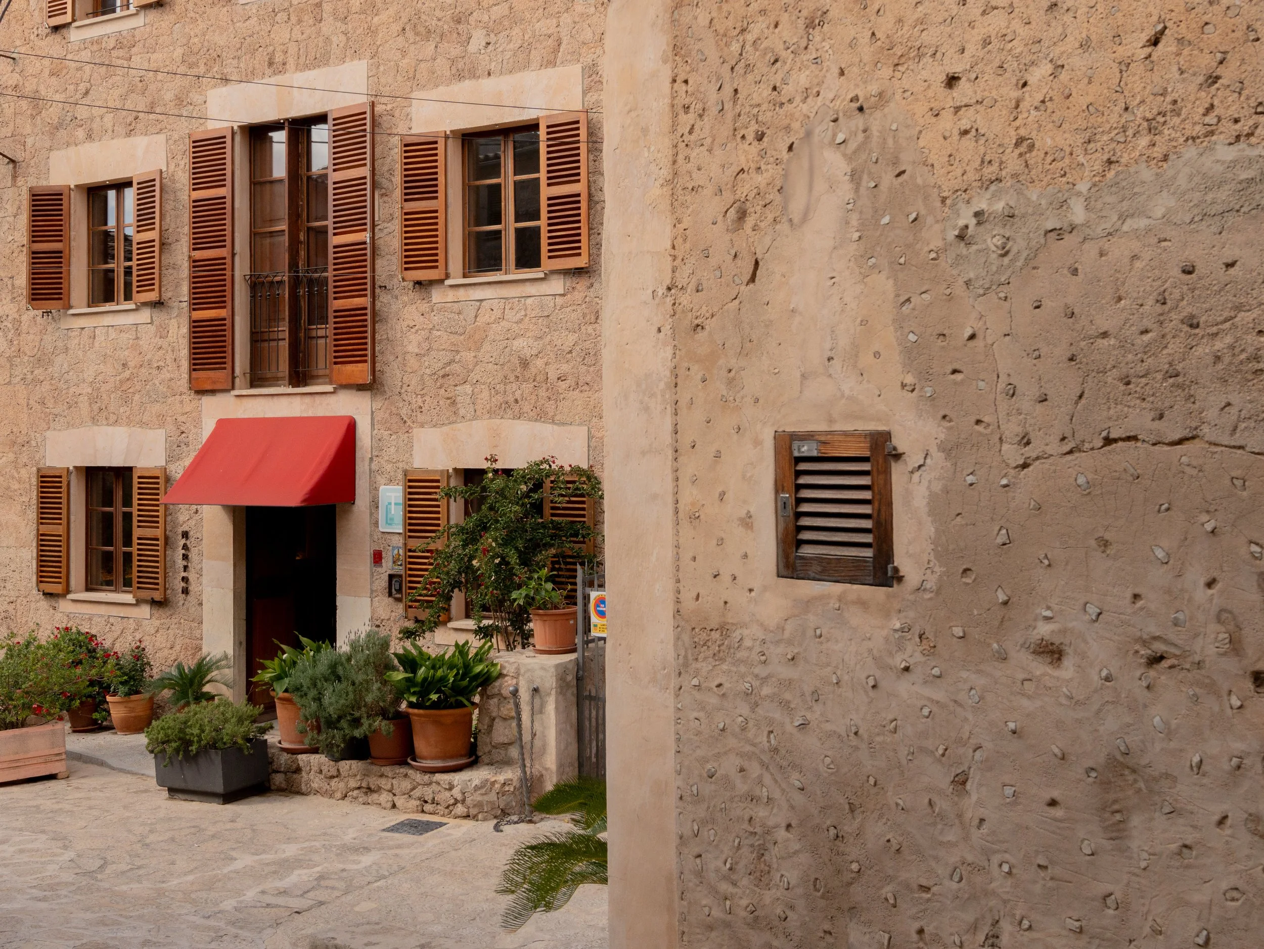 Casa con paredes de piedra, ventanas con persianas de madera y una entrada con toldo rojo, rodeada de macetas con plantas.