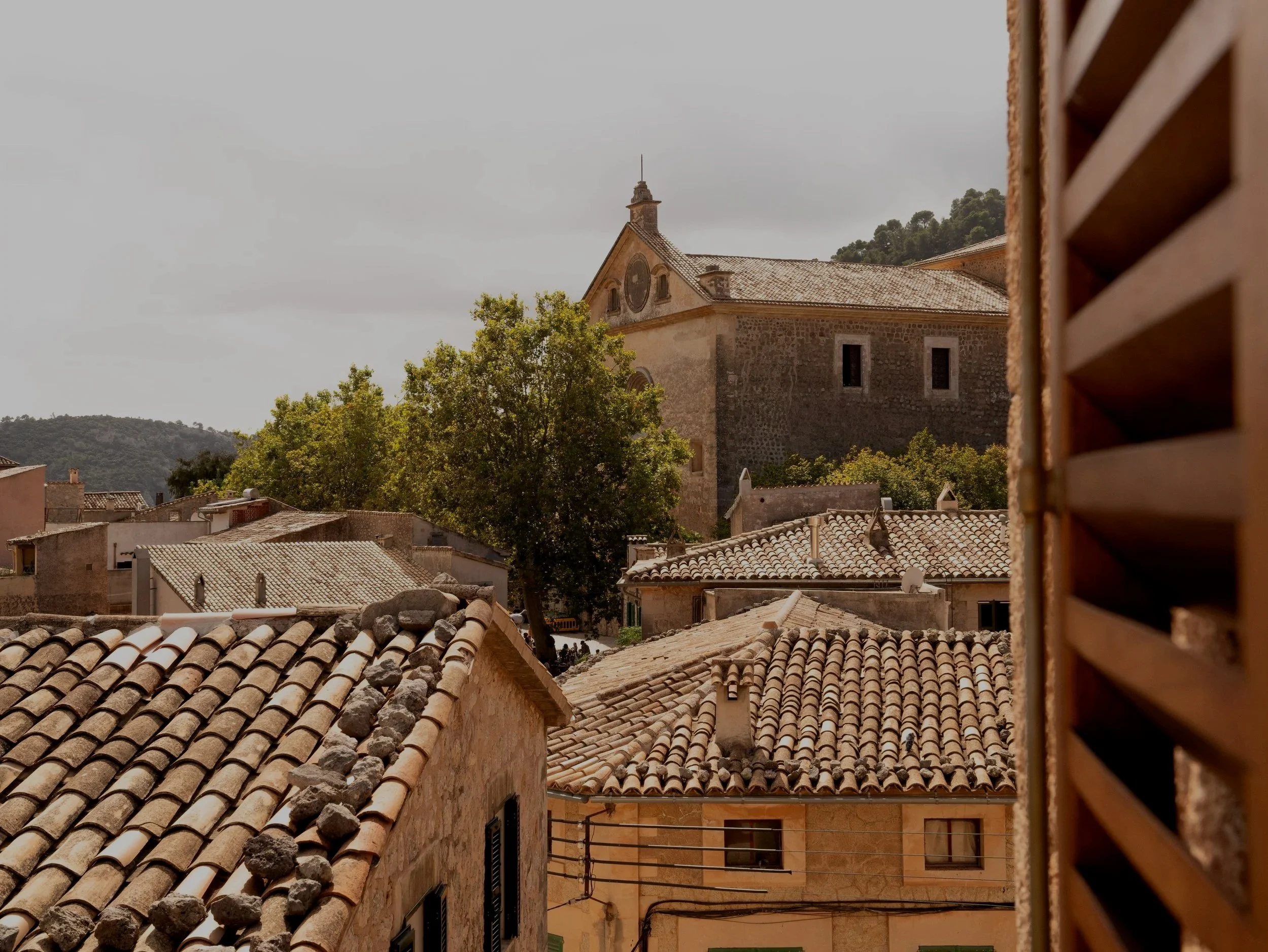 Vista de un pueblo con techos de tejas y un edificio grande en el fondo, árbol y montañas al fondo, vista desde una ventana con celosía de madera en el lado derecho