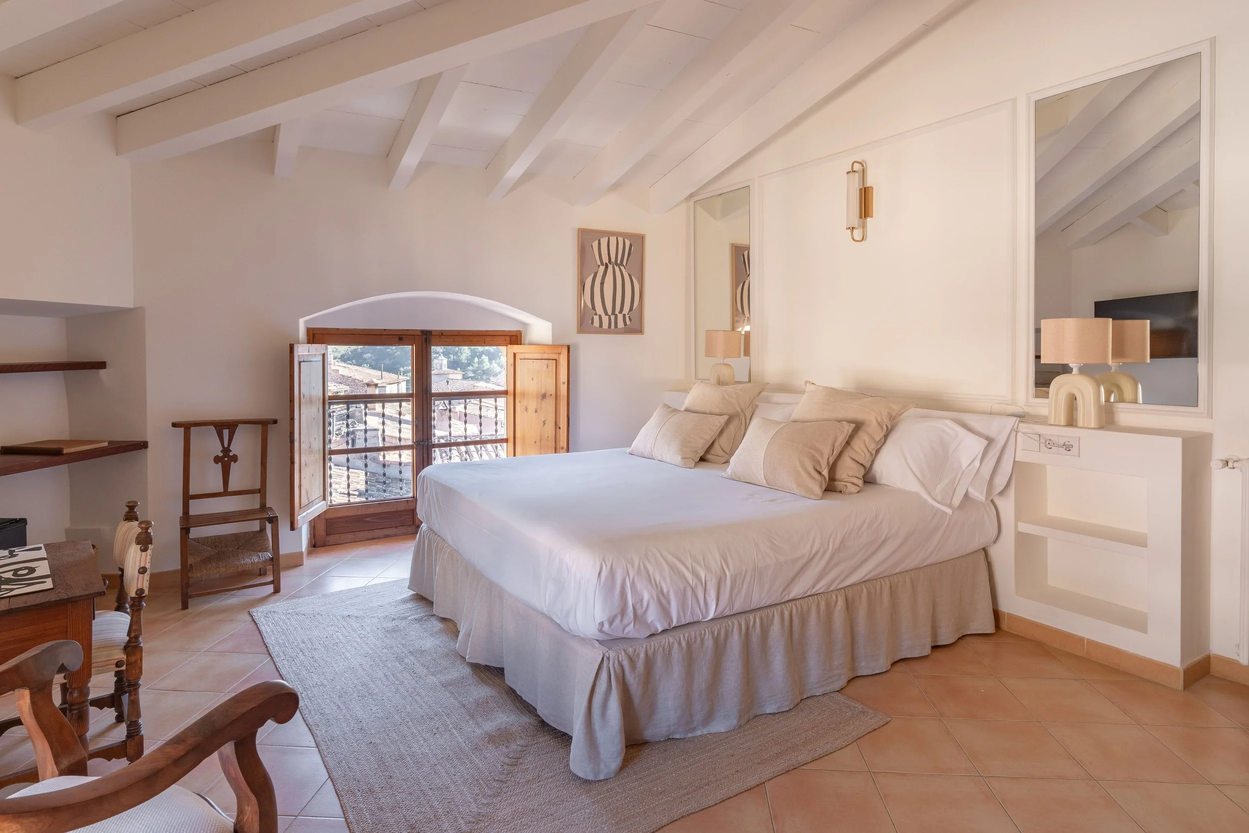 Habitación con cama grande, almohadas beige, ventanas de madera con vista al exterior, paredes blancas, lámparas, arte decorativo y pisos de baldosas.