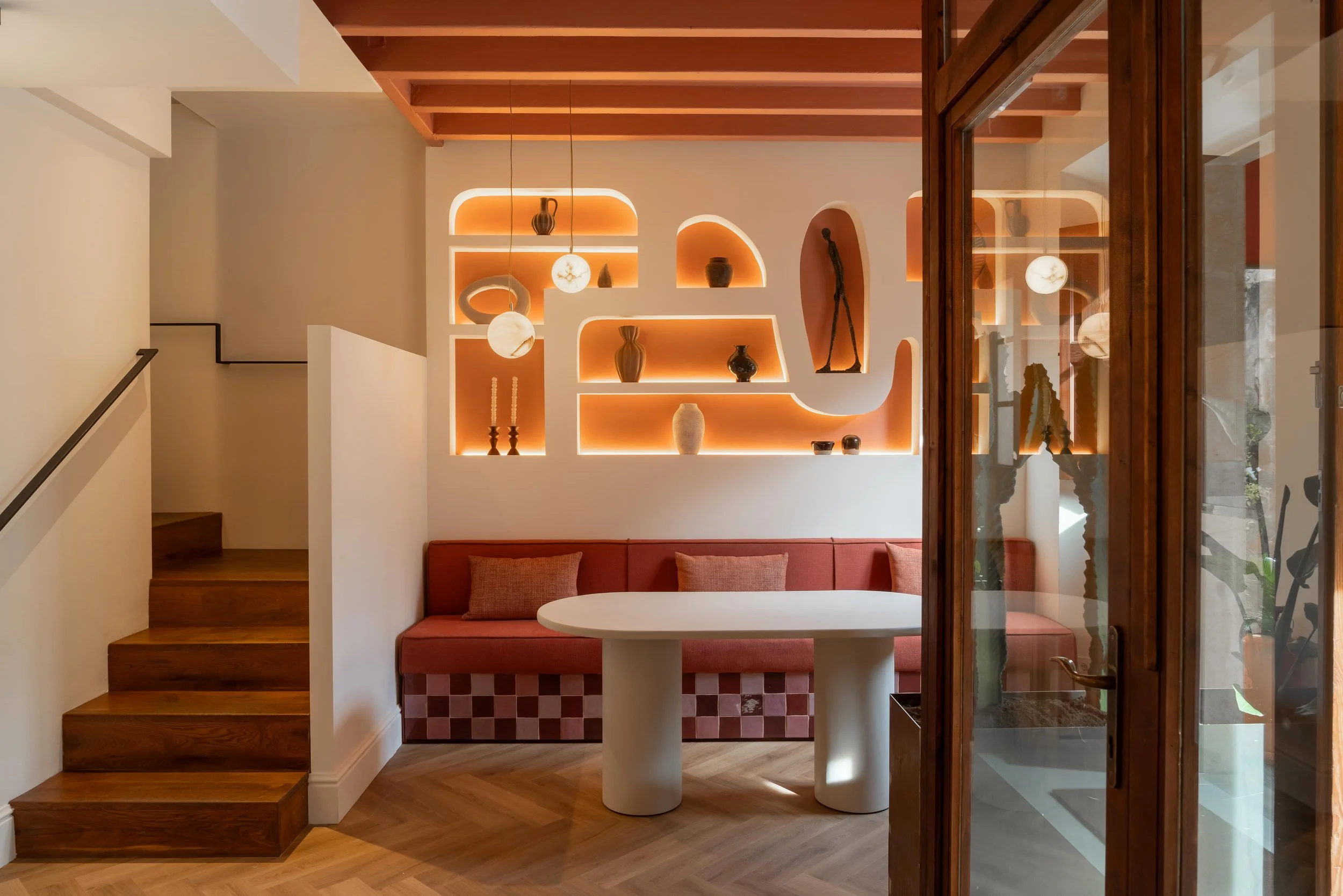 Interior acogedor con estantería de nichos con iluminación cálida, sofá de esquina con cojines, mesa redonda blanca, escaleras de madera y puerta de cristal con madera.