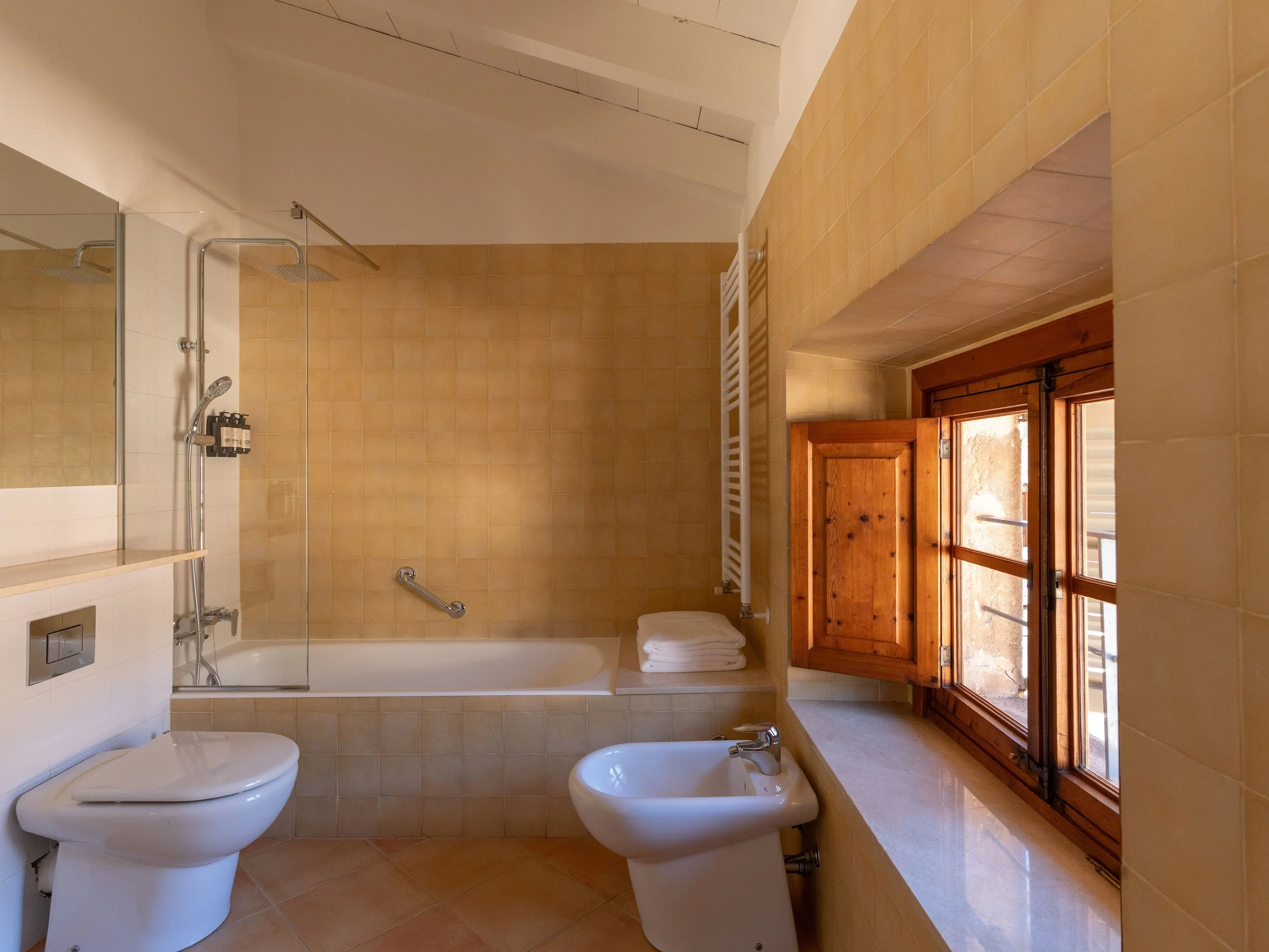 Baño con lavabo, WC, ducha, bidé, ventanas de madera y azulejos beige.