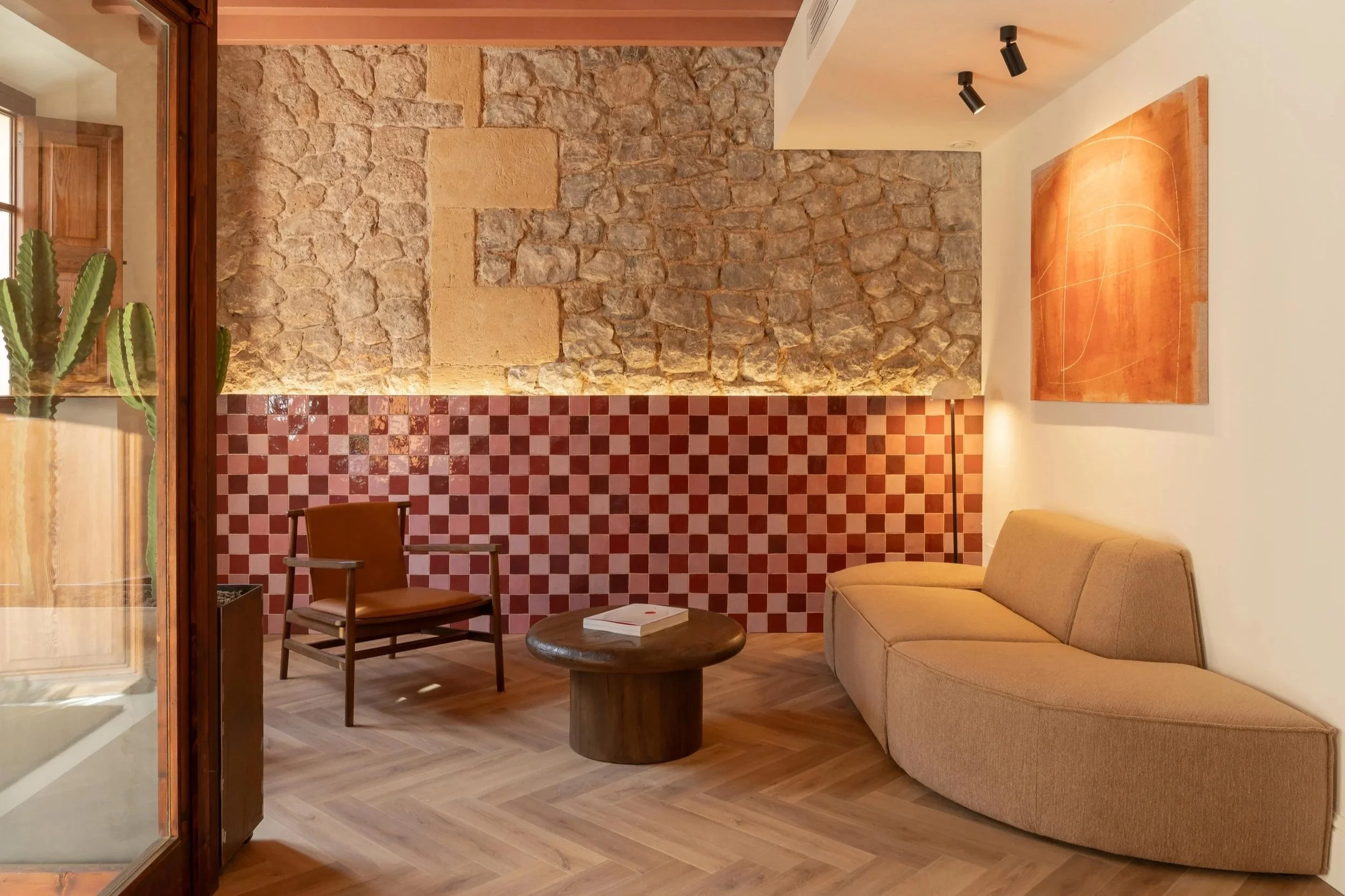 Sala de estar con sofá beige, silla de madera con cojín, mesa redonda de madera, pared con cuadros de piedra y azulejos rojos y blancos, planta cactus, y pintura moderna en la pared.