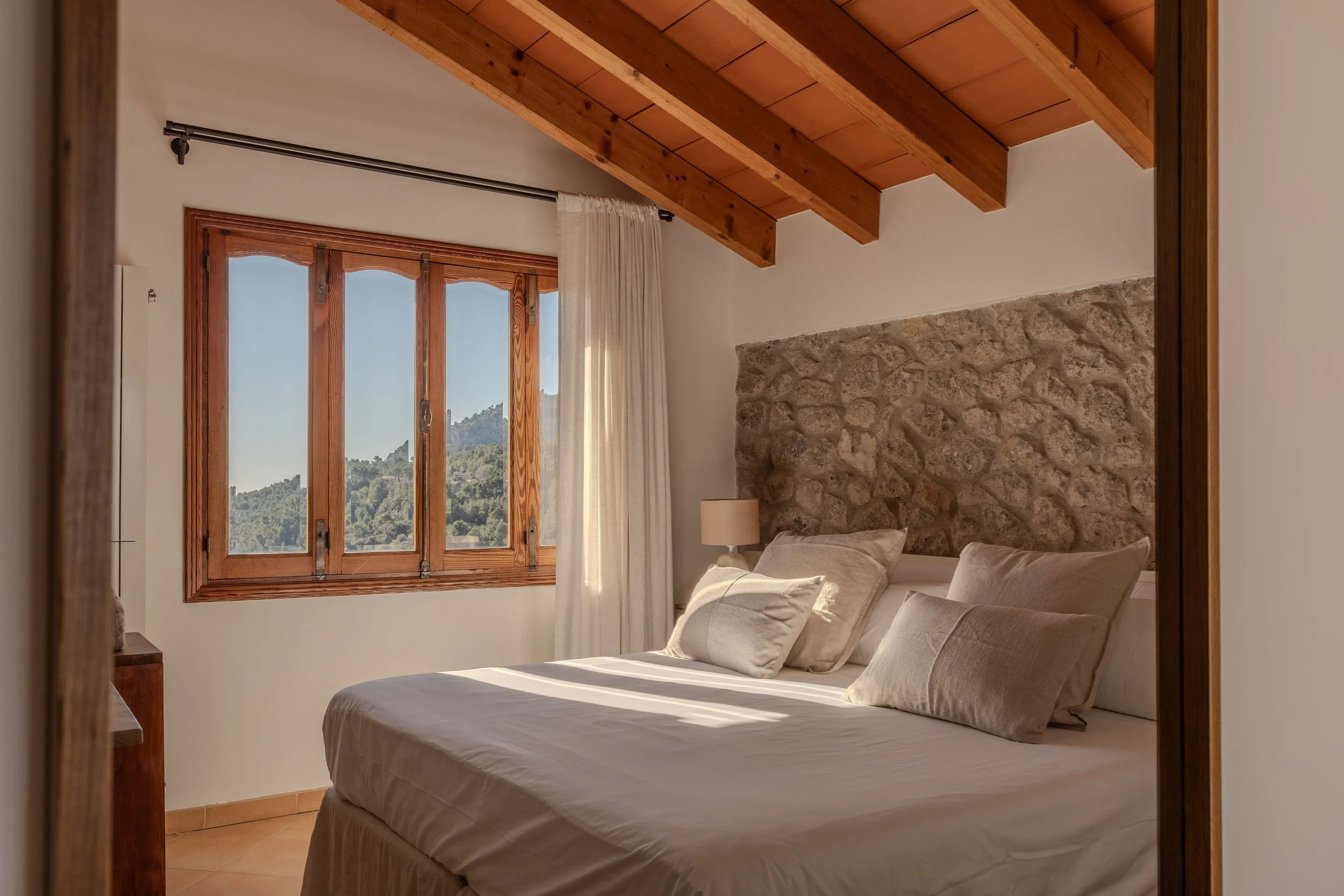 Habitación con cama, almohadas, lámpara y ventana con vista a montañas.