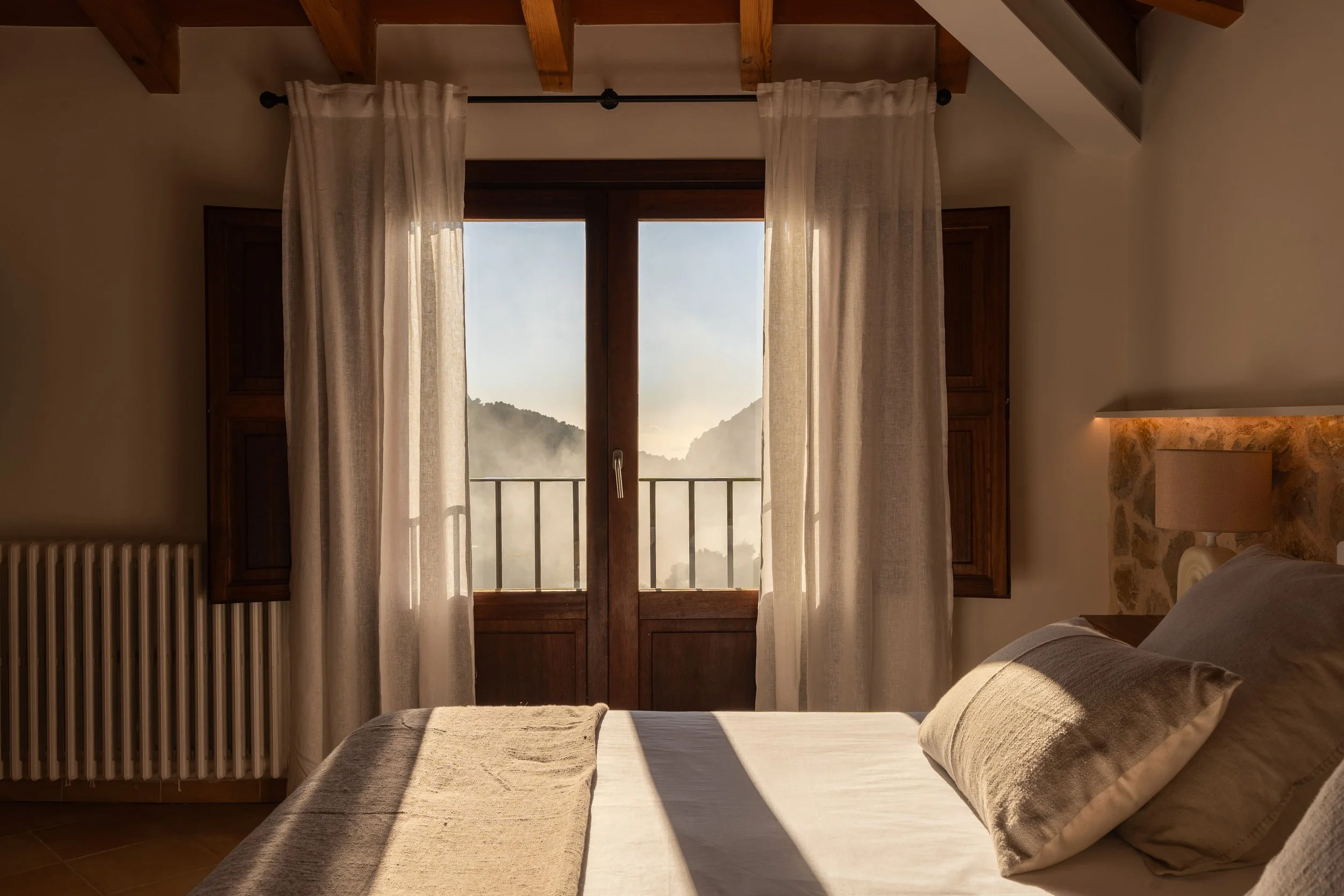 Habitación con cama y vista a las montañas a través de puertas de madera y cortinas claras.