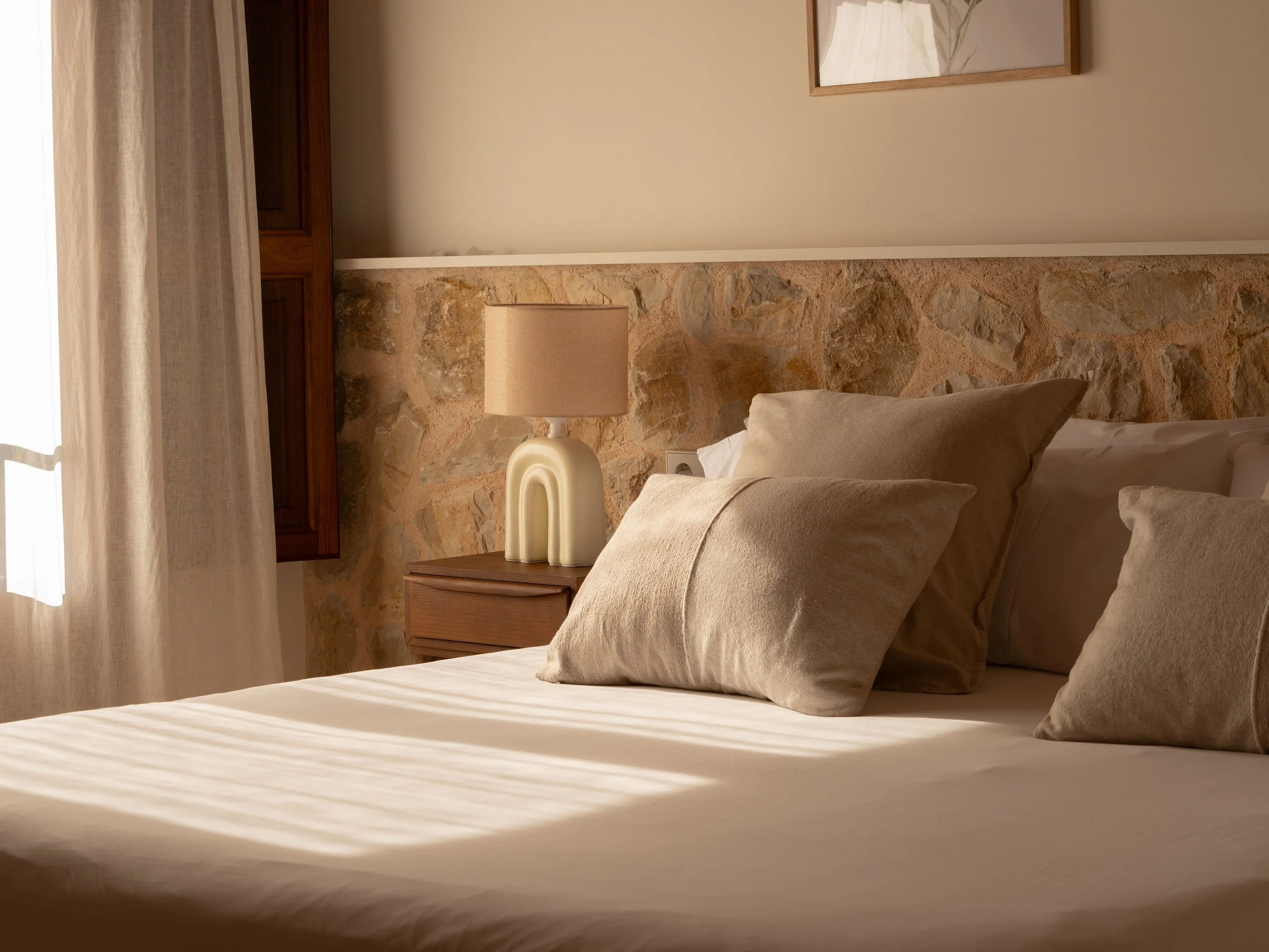 Cama con almohadas beige y lámpara sobre mesita de noche en habitación con pared de piedra y cortinas claras.