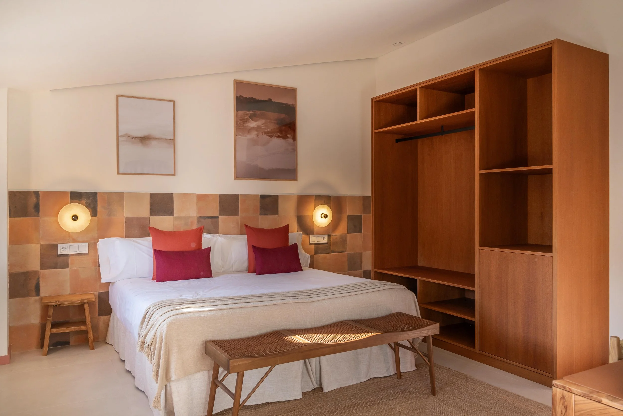 Habitación con cama grande, almohadas rojas y decoraciones de tonos cálidos, lámparas y arte en las paredes, armario de madera y mesitas laterales.