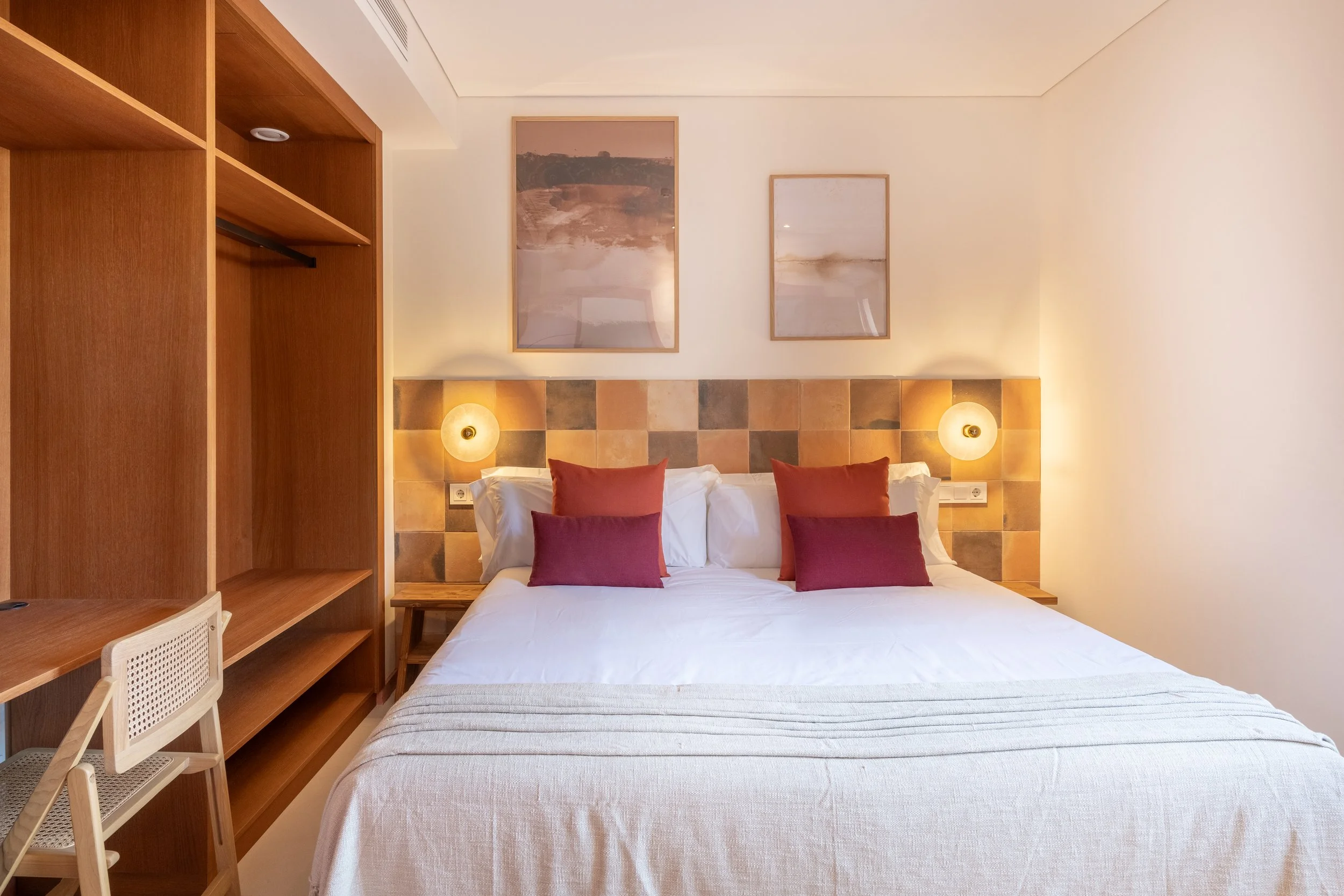 Habitación con cama grande, cabecera de azulejos, iluminación con apliques de pared, cuadros abstractos en la pared y mueble de madera.