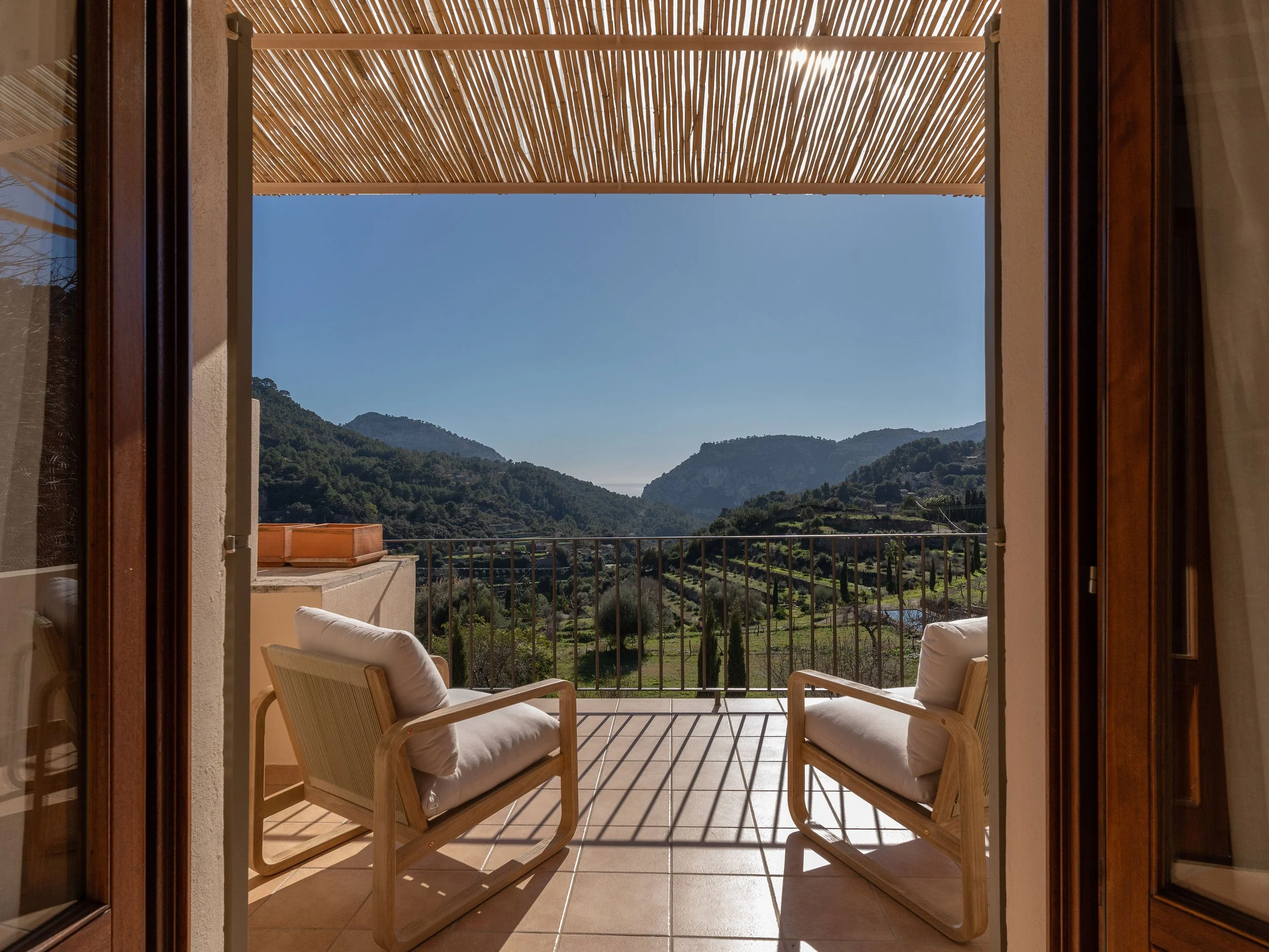Vista desde una terraza con dos sillones de madera y cojines blancos, vista de montañas y valle con vegetación, en un día soleado.