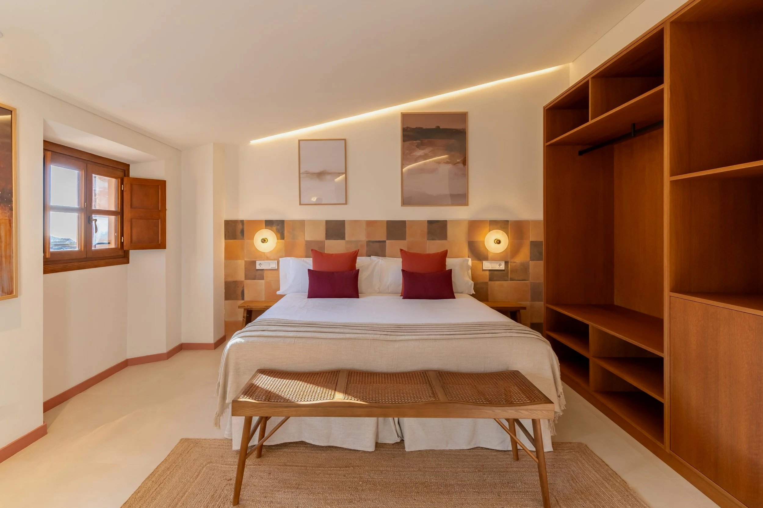Habitación con cama grande, cuatro almohadas rojas, paredes decoradas con cuadros y luces de pared, ventana con persianas de madera, muebles de madera clara, y una banca al pie de la cama, en un estilo rústico y acogedor.