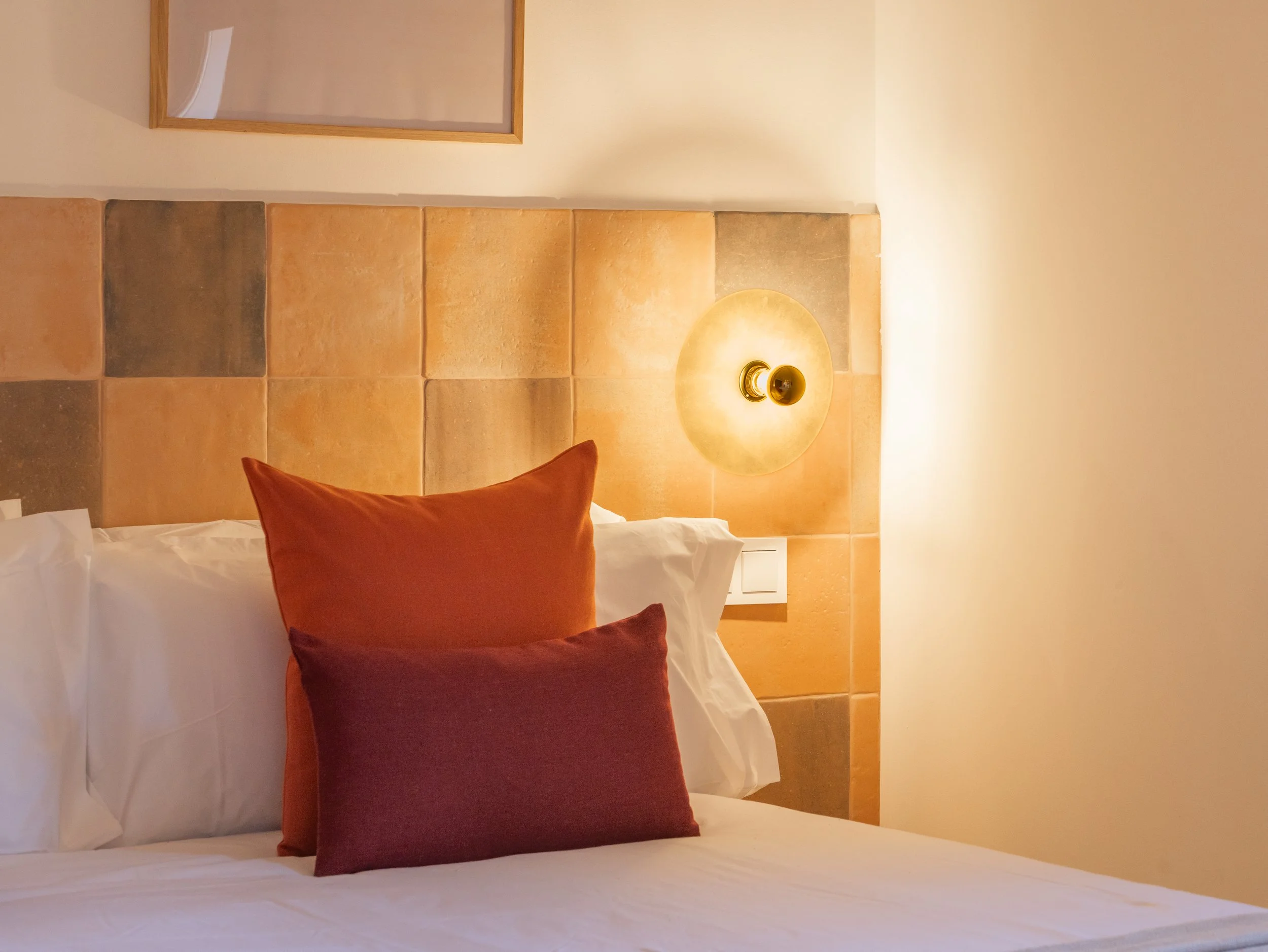 Cama con almohadas naranjas y color vino en una habitación con pared de azulejos y lámpara de luz cálida.