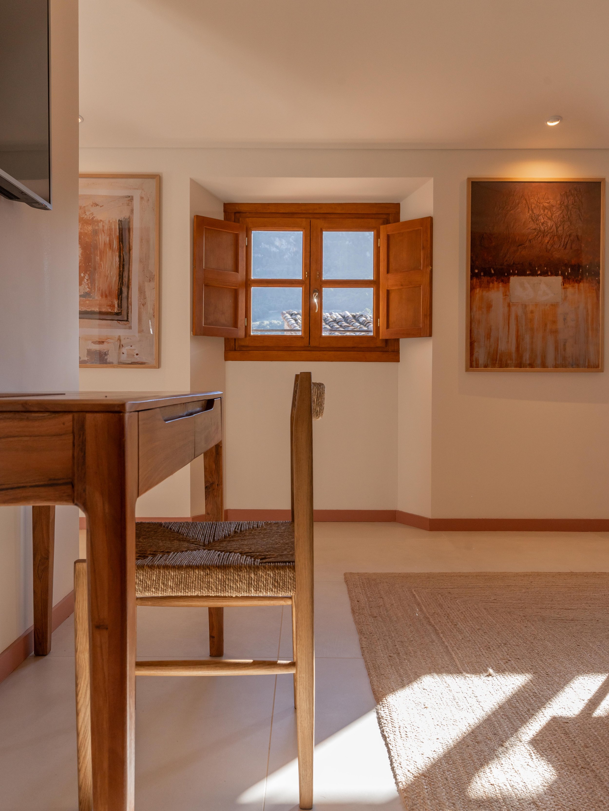 Habitación con paredes blancas, ventana de madera con postigos abiertos, cuadros abstractos en las paredes, alfombra de yute y mueble de madera.