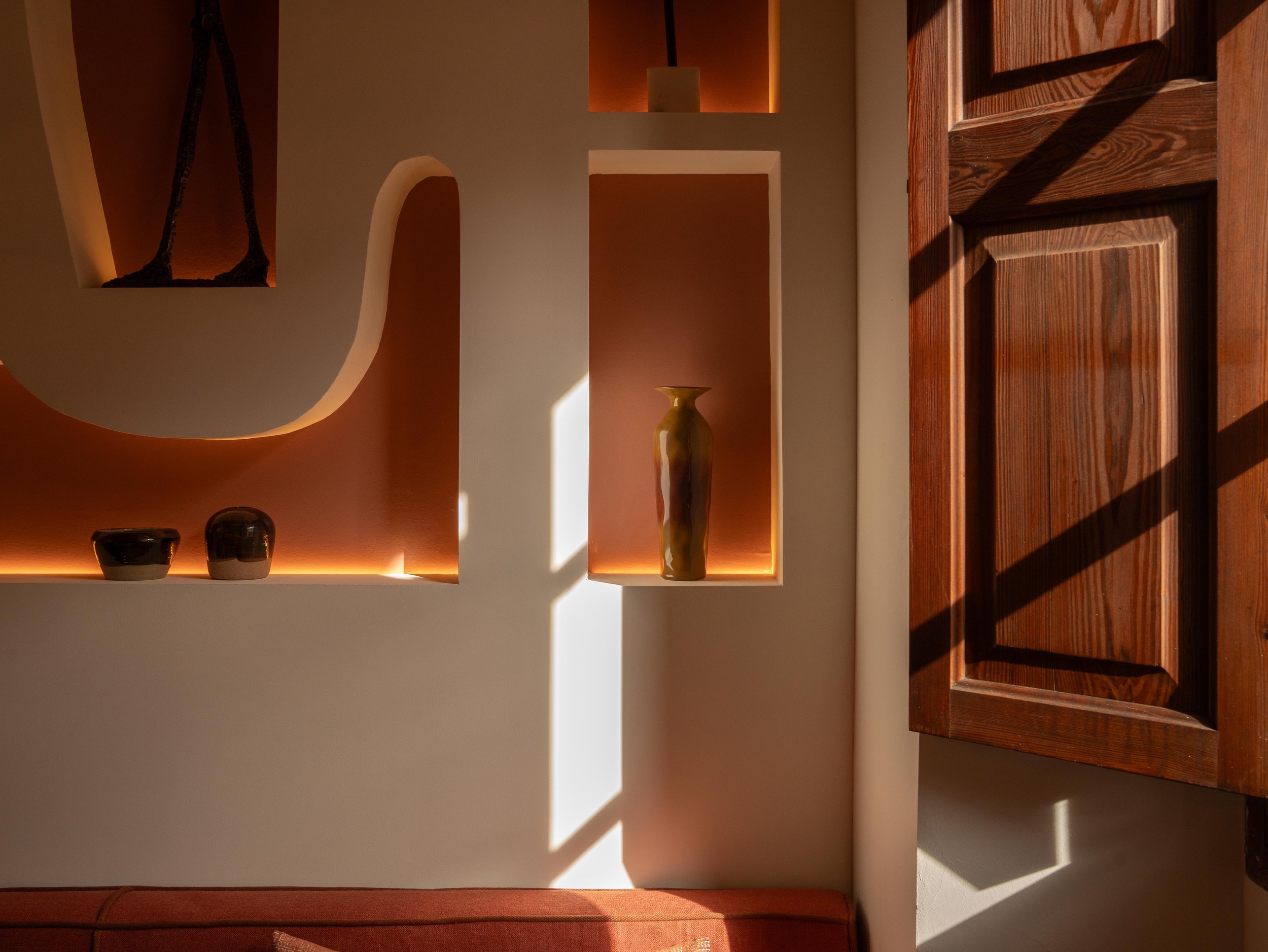 Decorative interior wall with built-in niches, illuminated with warm lighting, displaying vases and bowls, next to a wooden door with sunlight and shadow patterns.
